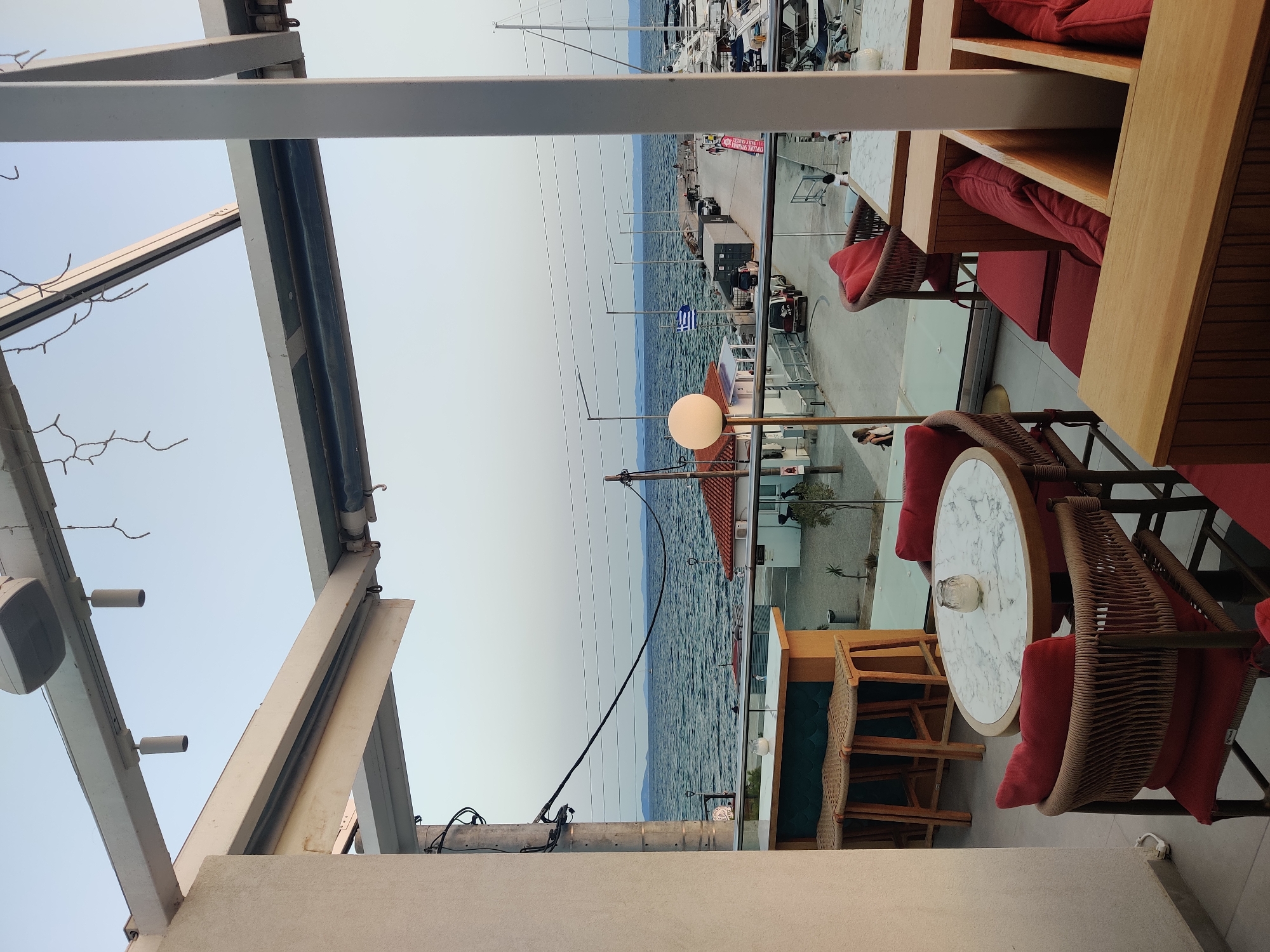 The image shows an outdoor seating area of a cafe or restaurant with a view of the sea and a harbor in Sithonia Municipal Unit, Greece. The seating area is furnished with wooden tables and chairs with red cushions. In the foreground, there is a marble-topped round table with chairs. Behind it, a bar with stools is visible. The background features a body of water, likely the Aegean Sea, with boats docked in a harbor. A pier with people walking and cars is also visible. The sky is clear and pale blue, suggesting it might be early morning or late afternoon. The overall atmosphere is relaxed and seaside-oriented.