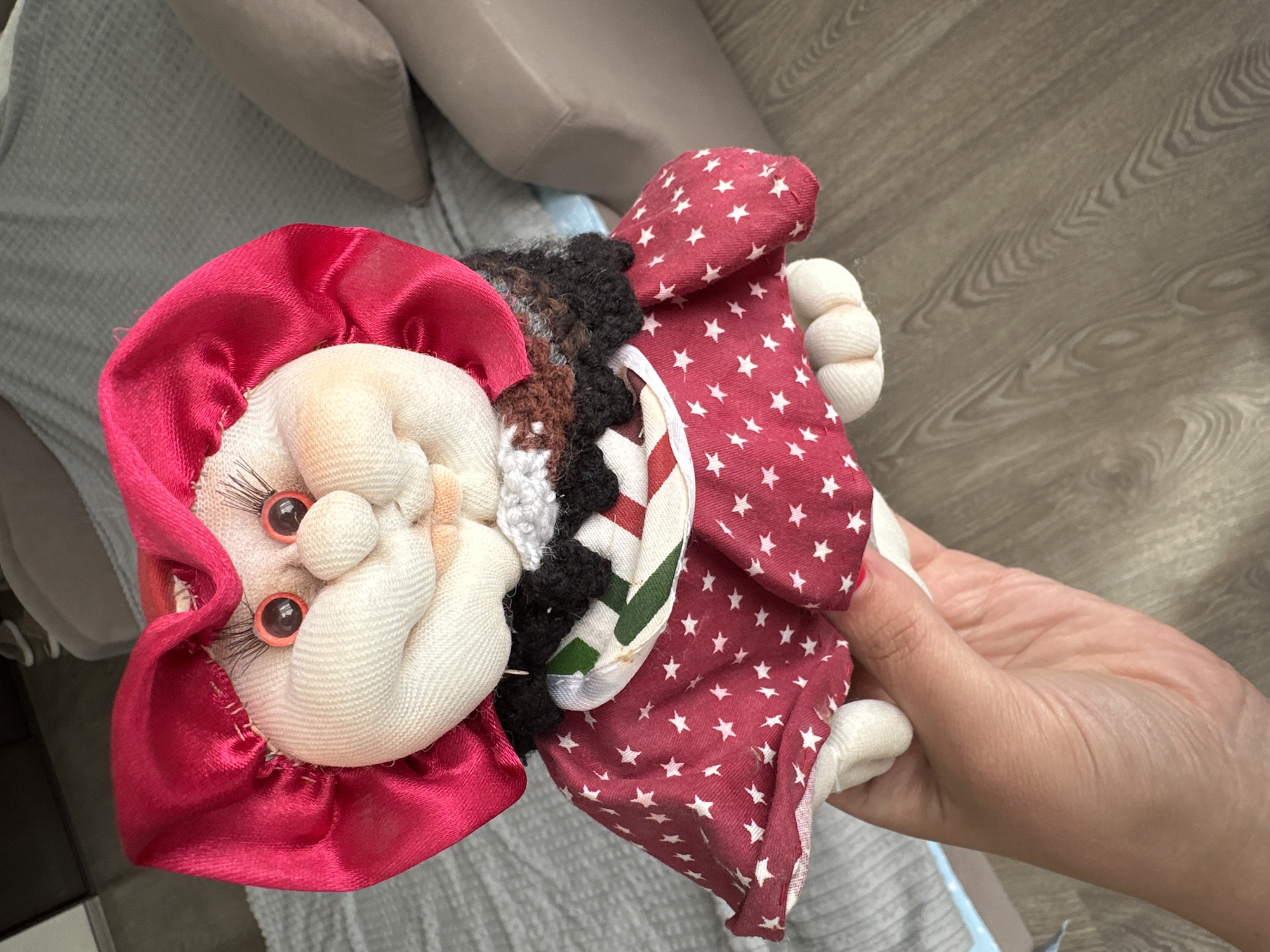 A handmade doll with a red bonnet and a red dress with white stars is held by a person's hand. The doll has round plastic eyes, a bulbous nose, and a smirking mouth. It is wearing a black and white ruffled collar. The background is blurred, showing a grey sofa and a wooden floor. The image is taken indoors, in what appears to be a residential setting. No specific location in Lytkarino, Russia, can be determined from the image.