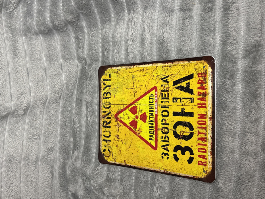 A rectangular, aged, yellow sign with a distressed finish lies on a soft, textured gray surface. The sign features the word "CHORNOBYL" vertically on the left. A red radiation hazard symbol sits above Ukrainian text and the words "ЗАБОРОНЕHA ЗОHA" in large, bold letters. The words "RADIATION HAZARD" are displayed on the right side. The edges of the sign are corroded.