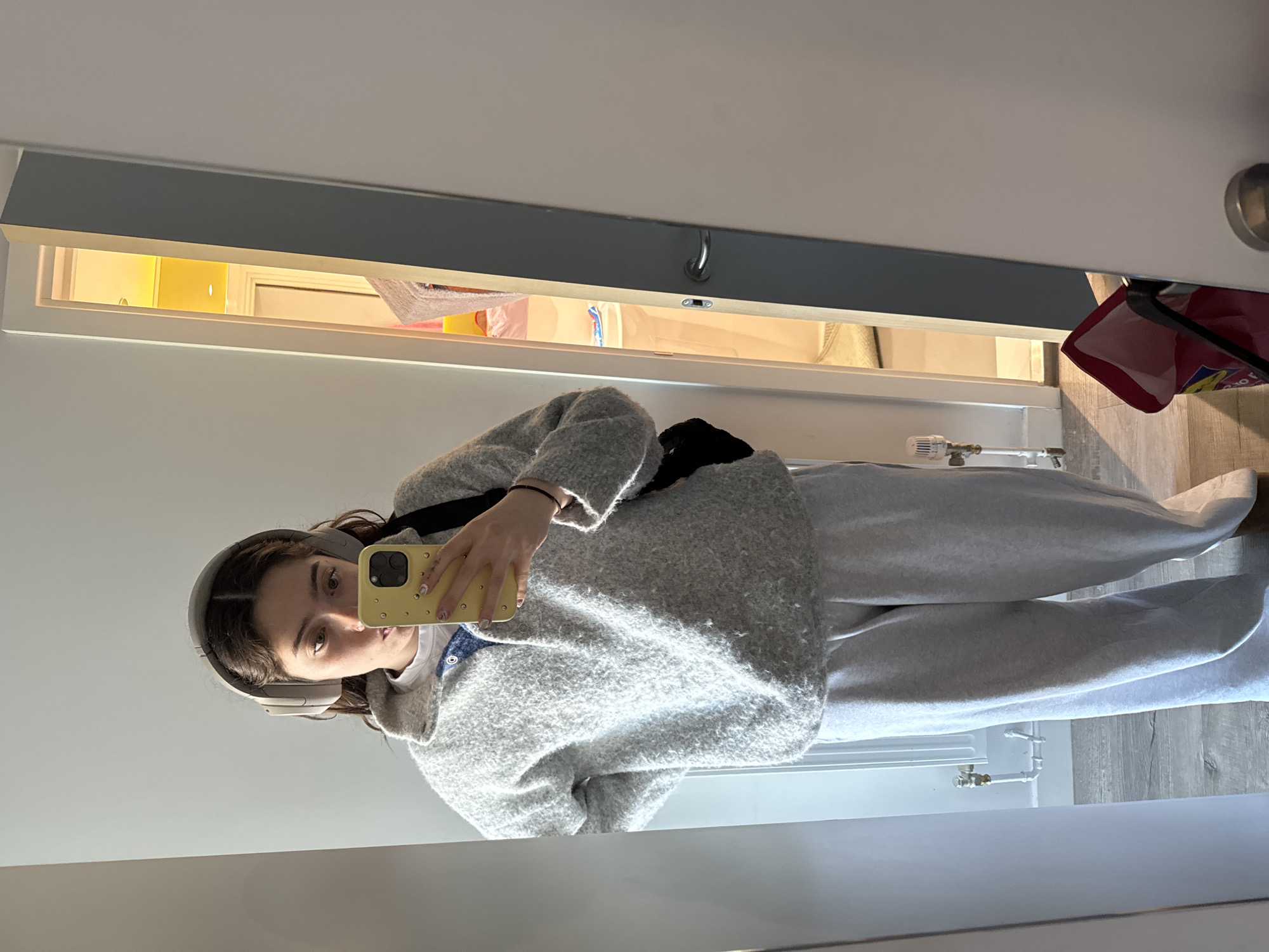 A young woman is captured in a mirror selfie, holding a yellow-cased smartphone in her right hand to take the photo. She has dark hair, wears light grey over-ear headphones, and is dressed in a light grey textured cardigan or jacket over a white t-shirt, paired with matching light grey sweatpants. A black strap from what appears to be a fanny pack or crossbody bag is visible across her chest.
The setting is an indoor space, likely a hallway or bedroom. The floor is covered with light-toned wooden planks. In the bottom right corner of the mirror's reflection, a white radiator with brass piping and a valve is partially visible. Directly above the mirror, there's an open, lit storage compartment or shelf. This area emits a warm, yellow glow and contains various items, including what looks like white fabrics, yellow objects, and a red bag with the partial text "Ofo" visible. A small metal hook is also attached to the underside of the shelf's frame. The walls are plain white. The overall impression is a casual, domestic scene, typical of a home in the United Kingdom. The bright lighting suggests it is daytime.