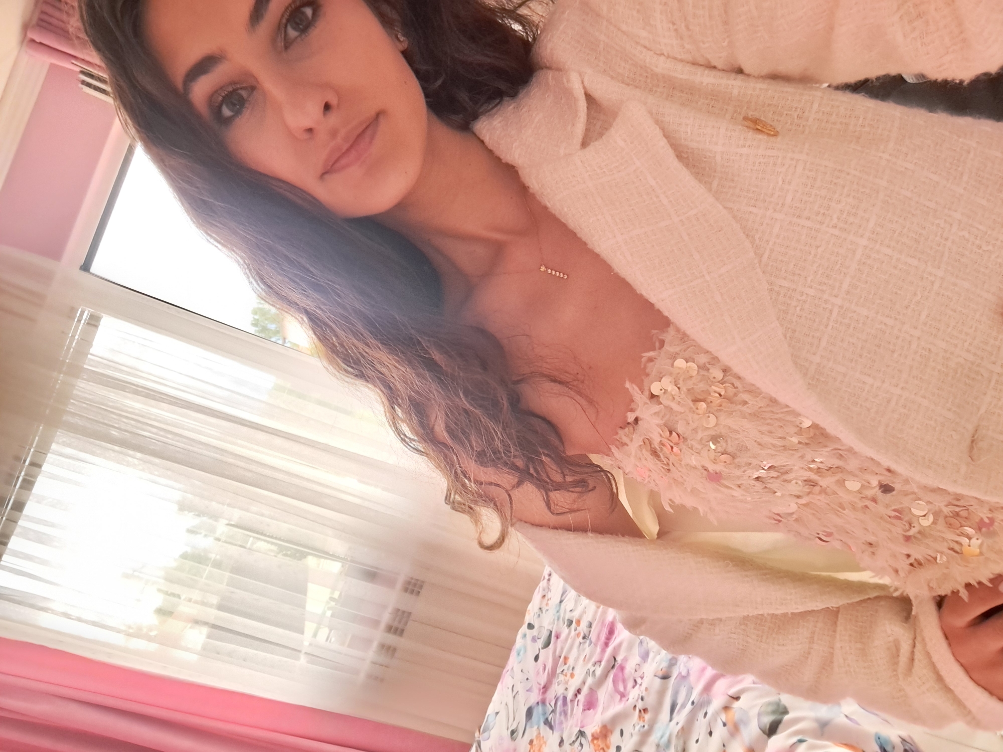 In Negovan, Bulgaria, a young woman with long, dark, wavy hair is captured in a close-up, high-angle shot. She is wearing a light-colored, textured blazer over a sequined, feathery top. A delicate necklace with a charm is visible around her neck. Her makeup includes noticeable eyeliner and eyeshadow. She is positioned in front of a window with sheer white curtains, partially obscured by a pink curtain. Natural light streams in from the window, illuminating the scene. In the background, a radiator is visible on the wall, and part of a bed with a floral-patterned comforter is also in frame. The overall impression is one of indoor comfort, possibly a bedroom, with soft lighting and feminine decor.