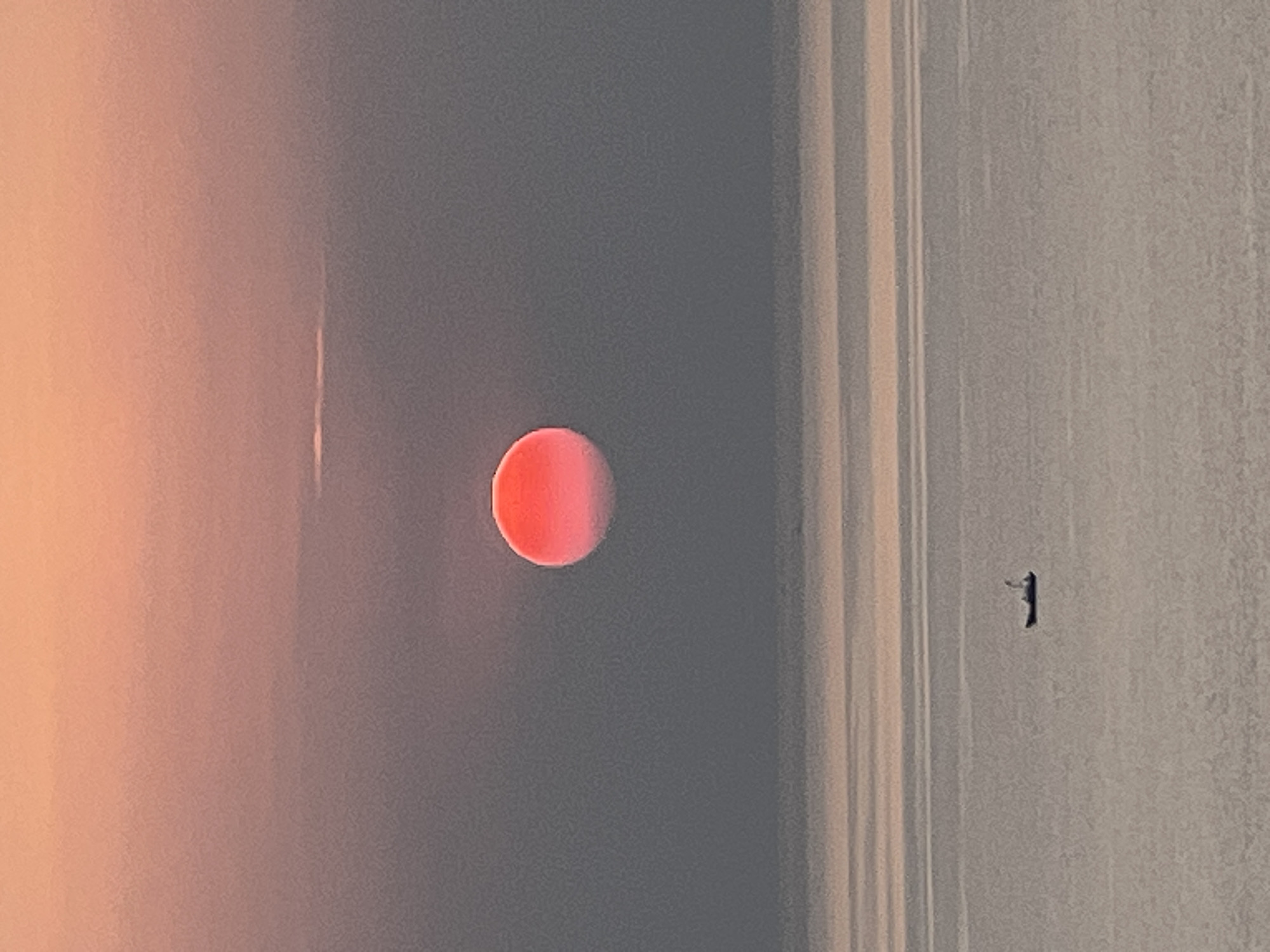 A bright orange sun is partially obscured by a hazy sky, casting a soft glow over a calm sea. The sky transitions from a soft peach hue at the top to a muted gray, with faint horizontal cloud formations visible. The sea is a tranquil expanse of light gray, with subtle ripples reflecting the dim light. A small, dark boat with a white sail is visible in the distance, floating on the water. The scene suggests a peaceful, possibly early morning or late evening, atmosphere. This image captures a moment of serene natural beauty along the coast of Nha Trang, Vietnam, known for its picturesque beaches and tranquil waters.