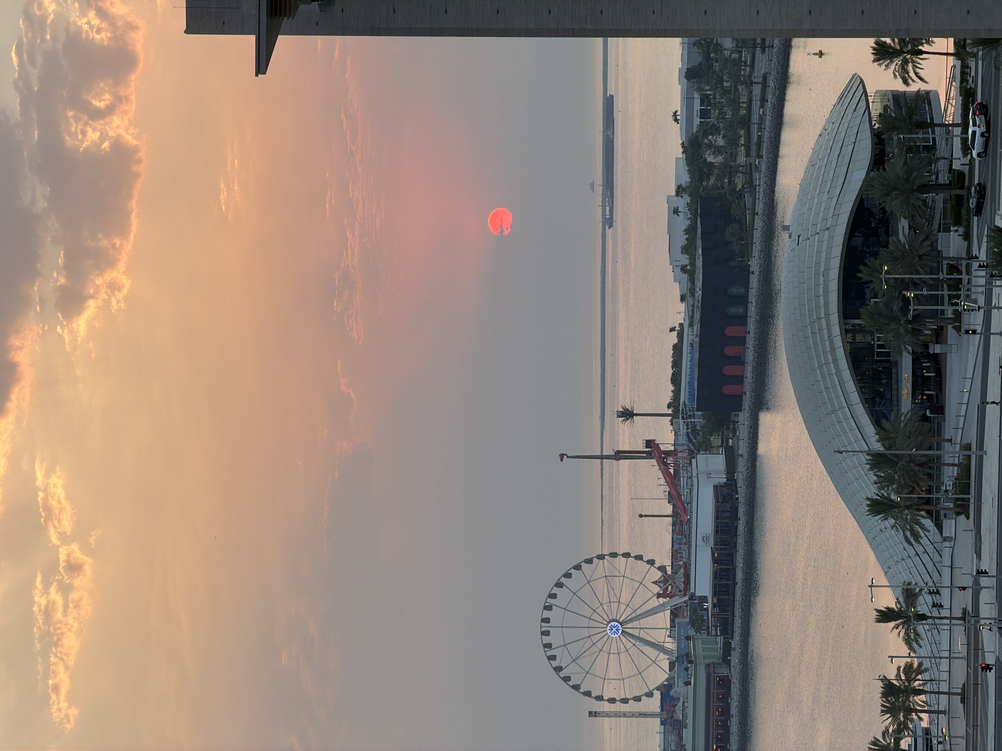 This is an aerial shot of the waterfront in Lusail, Qatar during sunset. The sky is filled with warm hues of orange and pink, with scattered clouds catching the fading sunlight. In the foreground, a modern building with a curved, metallic roof is prominent, surrounded by palm trees. Beyond this structure, a bay or canal separates it from an amusement park area. This area features a large Ferris wheel with illuminated spokes, several smaller rides, and various buildings that appear to house shops or restaurants. A body of water stretches to the horizon, where a large cargo ship and a smaller vessel are visible. To the right edge of the frame, a tall, modern building with a stone facade partially obstructs the view. The scene suggests a vibrant urban environment situated by the sea.
