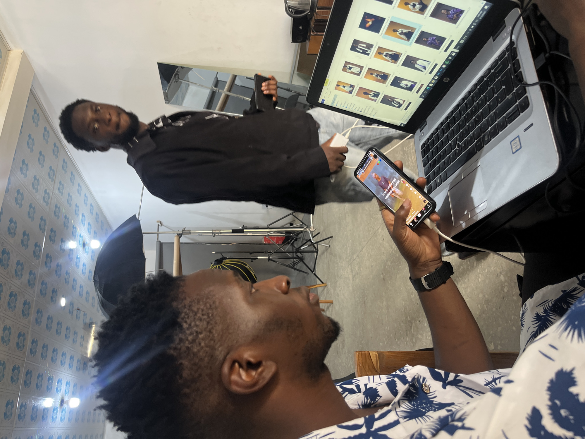 The image shows two men in what appears to be a photography studio. One man, facing the camera, is wearing a black jacket and looking towards a laptop. He is holding a smartphone in his left hand. The second man, in the foreground and facing away from the camera, is wearing a white shirt with a blue floral pattern. He is also holding a smartphone, which displays text and an image.
The laptop screen shows a grid of thumbnail images, possibly of photographs. The smartphone held by the man in the foreground displays text that reads "how to make your relationship to work."
Behind the man in the black jacket, there are photography studio lights and equipment, including light stands and a large umbrella. The background shows a tiled wall with a blue pattern and a grey floor. The overall impression is of a creative space where work is being done, possibly related to photography or content creation. The lighting suggests it could be daytime, though no specific time is discernible. The overall mood appears to be focused and professional.