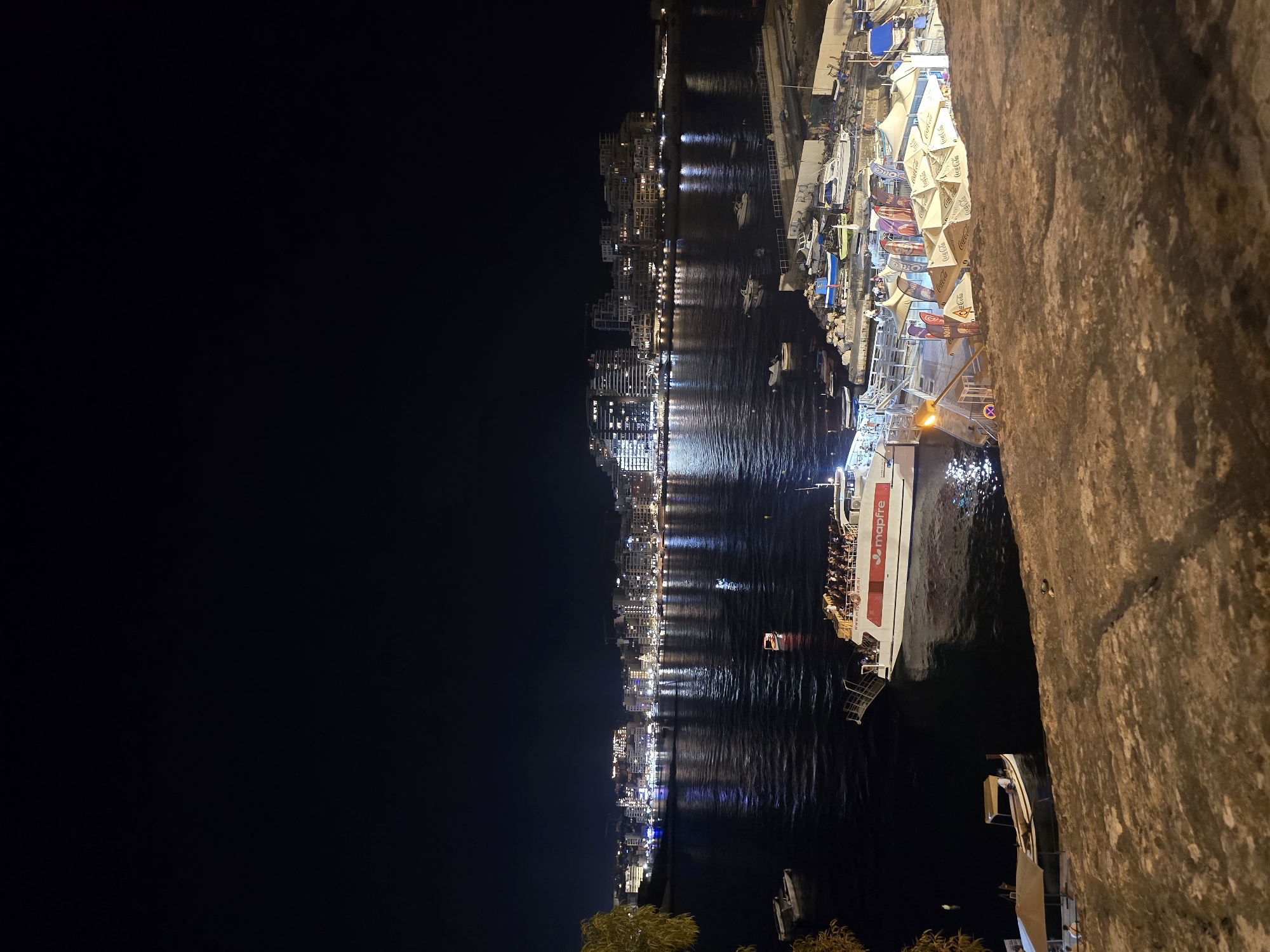 This is a nighttime aerial view of the Valletta waterfront in Malta. A large tour boat, the "Mapfre," is docked in the foreground, illuminated by its own lights and reflecting on the dark water. The boat is filled with passengers. Several smaller boats are moored nearby. Beyond the waterfront, the city of Valletta is visible, with its buildings illuminated by numerous lights, creating a bright glow against the dark sky. The water is calm, with gentle ripples reflecting the city lights. The foreground of the image is a textured stone wall, suggesting the vantage point is from a higher elevation, possibly within the fortifications of Valletta. A few trees are also visible on the left side of the frame. The scene depicts a bustling evening at a popular tourist and transportation hub in Valletta.