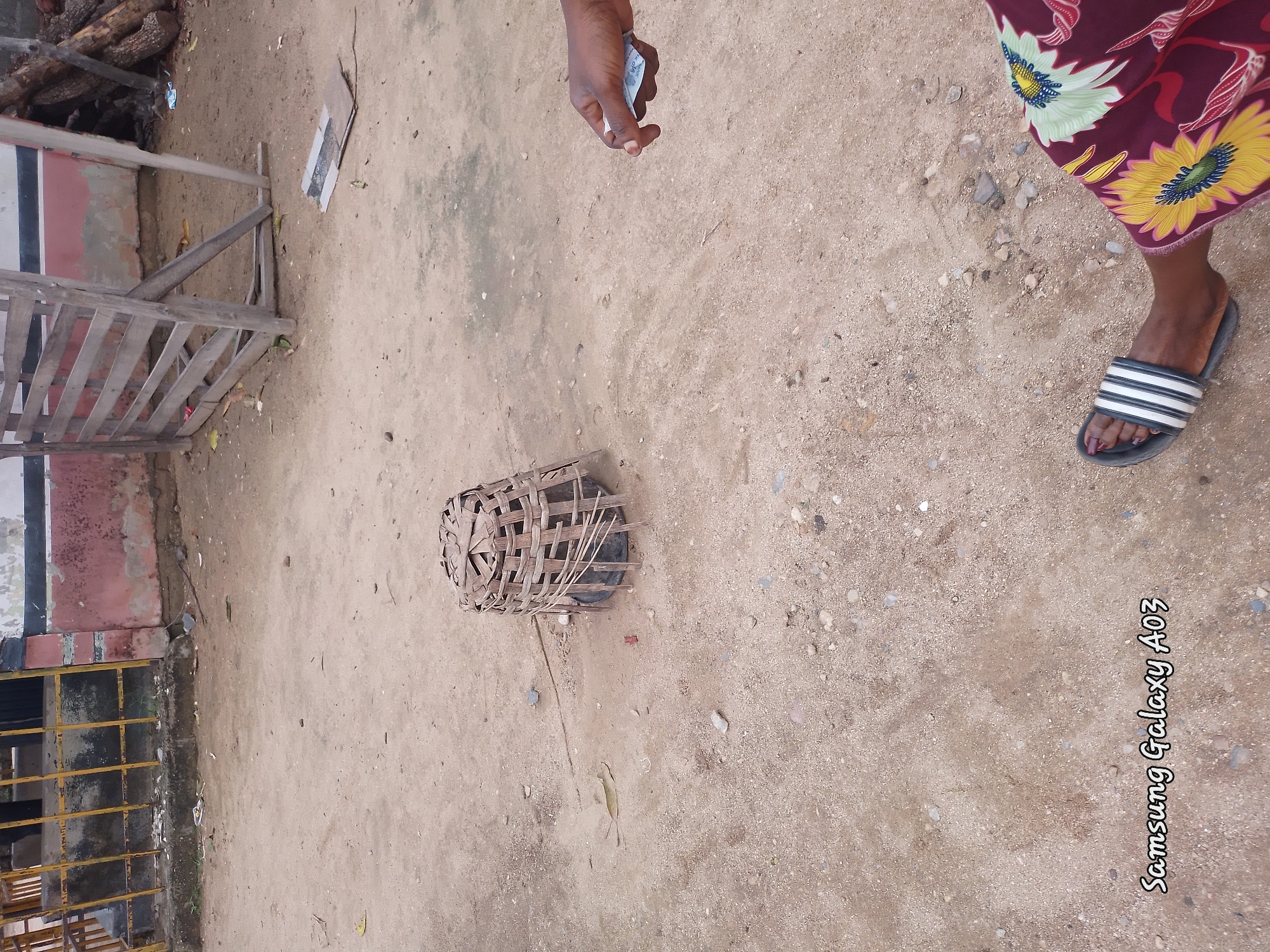 The image depicts an outdoor scene on a dirt ground, likely in Doka-Tofa, Nigeria. In the center of the frame, a woven basket, possibly a trap or container, sits on the ground. To the right, a person's hand is visible holding some currency. In the foreground, a person's foot wearing a striped sandal is visible, along with a portion of their floral-patterned skirt. The background shows a weathered wooden structure and some stacked logs, suggesting a rustic or possibly rural setting. A yellow metal fence is also visible on the left side of the image. The overall environment appears to be dusty and somewhat unkempt.