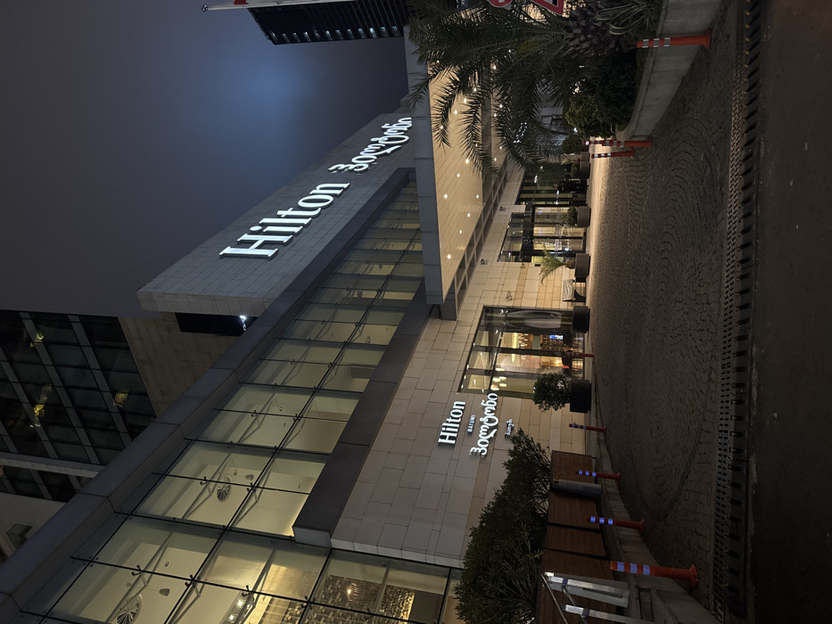 The image depicts the exterior of the Hilton Batumi hotel in Batumi, Georgia, at night. The modern, multi-story building features a light-colored lower facade with extensive glass paneling and a large, illuminated "Hilton" sign in white English letters and Georgian script ("ჰილტონ") positioned prominently on the upper section. A smaller "Hilton BATUMI" sign, also in English and Georgian ("ბათუმი"), is located above a ground-level entrance. A large, illuminated covered portico extends over a curved, cobblestone driveway that shows visible tire marks.
Two indistinct individuals are visible near the main entrance doors beneath the portico. The driveway is bordered by orange and white reflective traffic bollards. Planters containing shrubs line the left side of the driveway, while palm trees and other foliage are present on the right side, near the main entrance. A drainage grate is visible across the foreground of the driveway. The sky is dark and overcast. A flagpole with a red flag is visible on the building's roofline to the far right.