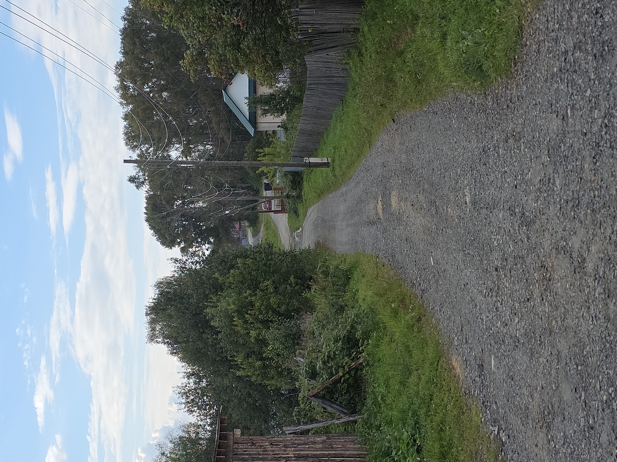 A gravel road winds through a rural area under a cloudy blue sky. On the left side of the road, there is a wooden structure and lush green vegetation. On the right, a weathered wooden fence borders a grassy area and a small house with a green roof. Power lines stretch across the scene, supported by utility poles. The road continues into the distance, flanked by trees and houses, suggesting it leads further into the settlement of Magnitka, Russia. There are no people or animals visible in the image.
