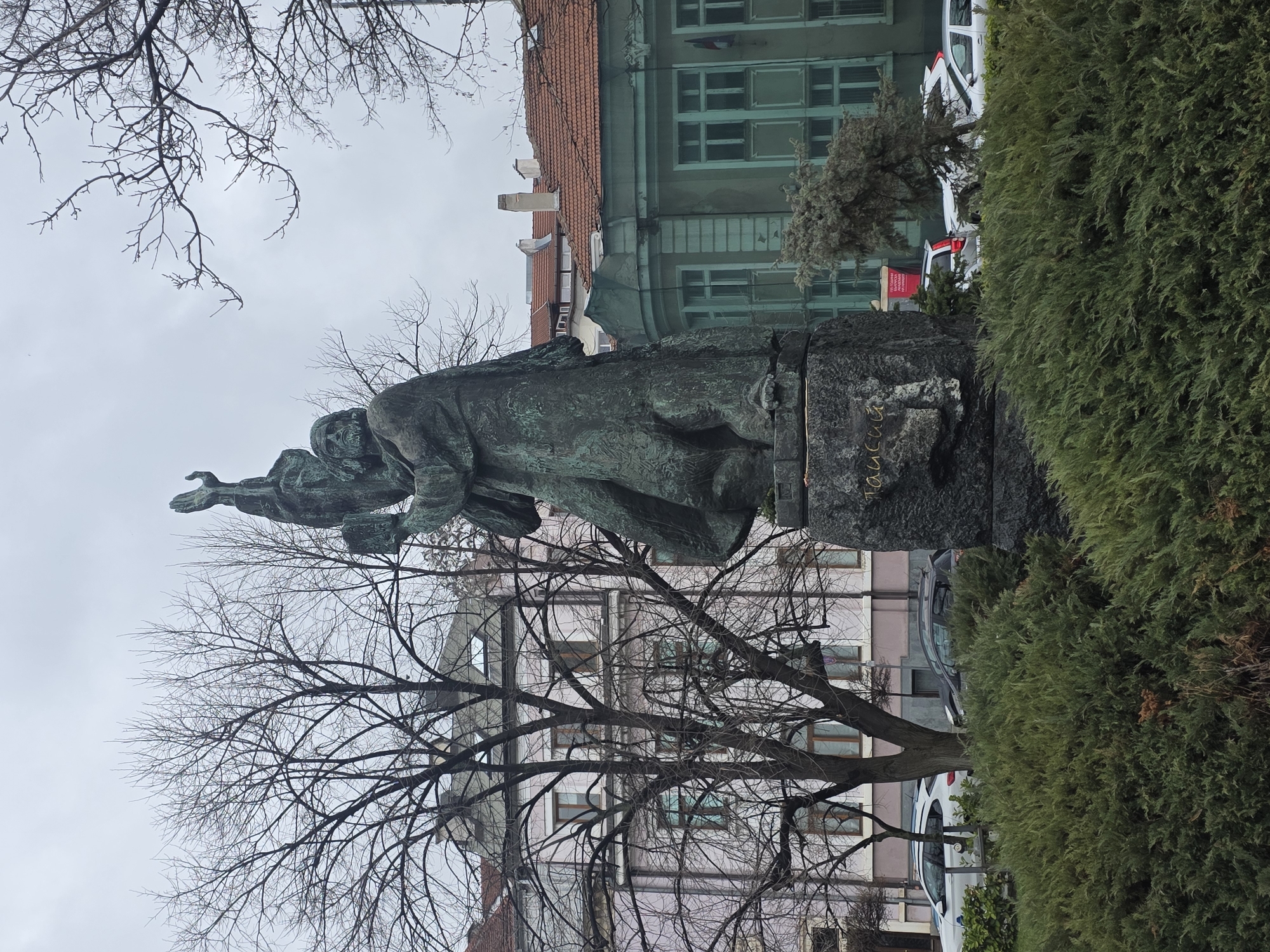 The image shows a statue of a kneeling figure in Sofia, Bulgaria. The statue is made of a dark-colored metal, possibly bronze or copper, and stands on a stone base. The statue depicts a man with his right arm raised in a gesture of supplication or offering. The statue is likely part of a monument or memorial. Behind the statue, there are buildings with windows, and bare trees with intricate branches. A tall, dense green hedge is visible on the right side of the frame. Parked cars are present in the background, indicating an urban setting. The sky is overcast.