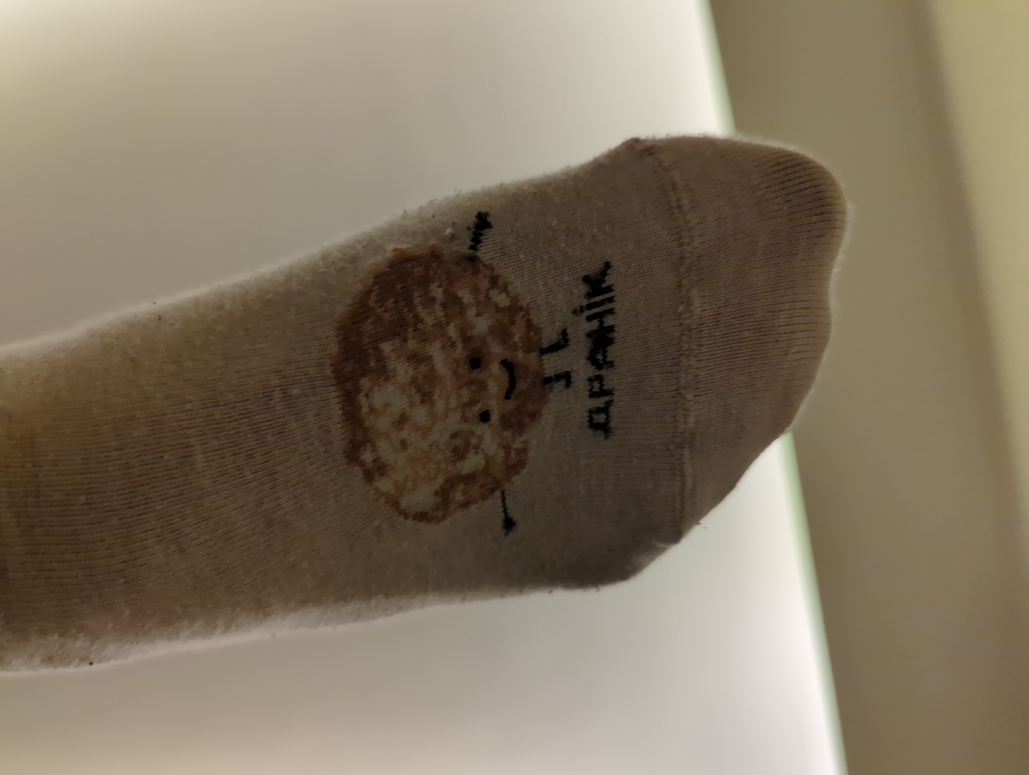 The image shows a close-up of a sock. The sock is light-colored and features a design of a round, smiling face resembling a pancake or pastry with small "legs" or antennae. Next to the design is the word "ДРАНІК" written in Cyrillic letters. Based on the design and the text on the sock, it is likely related to "draniki," which are potato pancakes popular in Belarusian cuisine. The image does not provide any information about the location of the photo being taken. Thus, it is unknown if it was taken in Minsk, Belarus, or anywhere else.