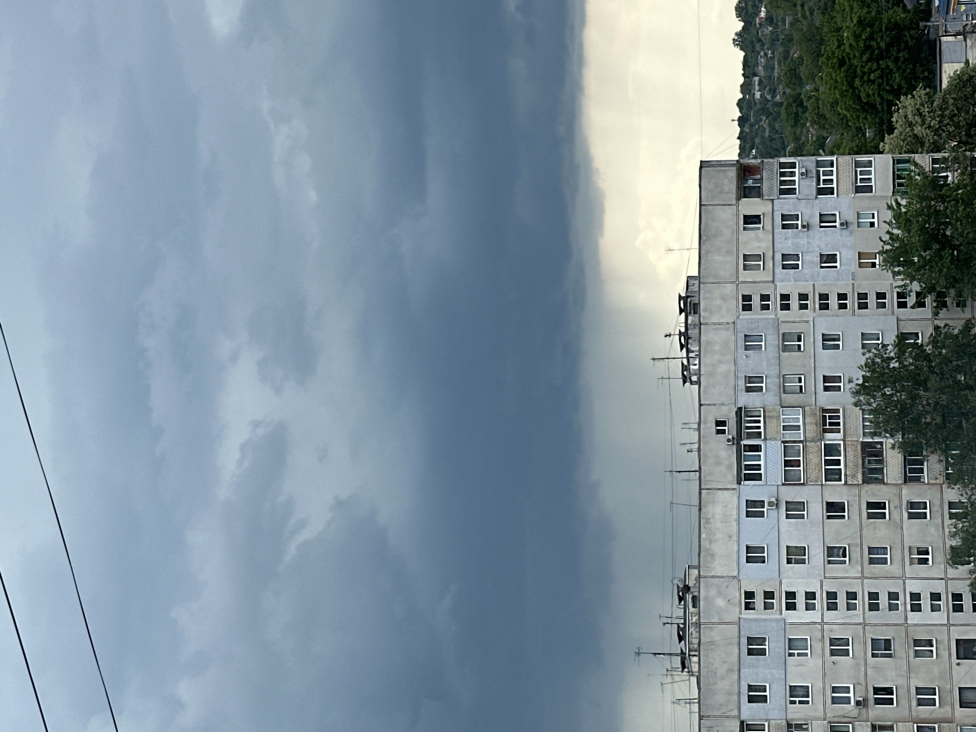 A vertically oriented photo shows a gray apartment building on the right and a stormy sky on the left. The sky is mostly dark gray, suggesting an approaching storm. A lighter gray area is visible to the right of the dark storm clouds, near the apartment building. The apartment building is a high-rise with many windows, and it appears to be an older construction. It is primarily gray in color, with individual rectangular window panes. Some trees are visible around the building, partially obscuring the view. Power lines are visible in the lower left portion of the frame, extending across the sky. The overall mood is somber, reflecting the stormy weather. A hill covered with green trees and possibly some structures is visible behind the apartment building.
