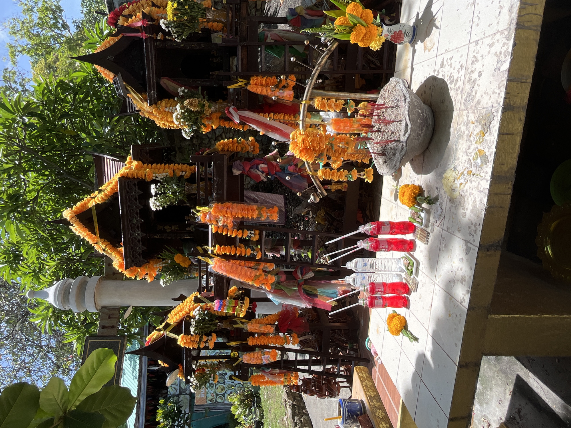 The image depicts a spirit house located in Baan Hua Thanon, Thailand. The spirit house is a multi-tiered structure adorned with orange and white flower garlands. Offerings of water bottles and flowers are placed on a tiled surface in front of the spirit house. An incense holder, filled with burning incense sticks, sits to the right of the structure. The scene is set outdoors with trees and greenery visible in the background.