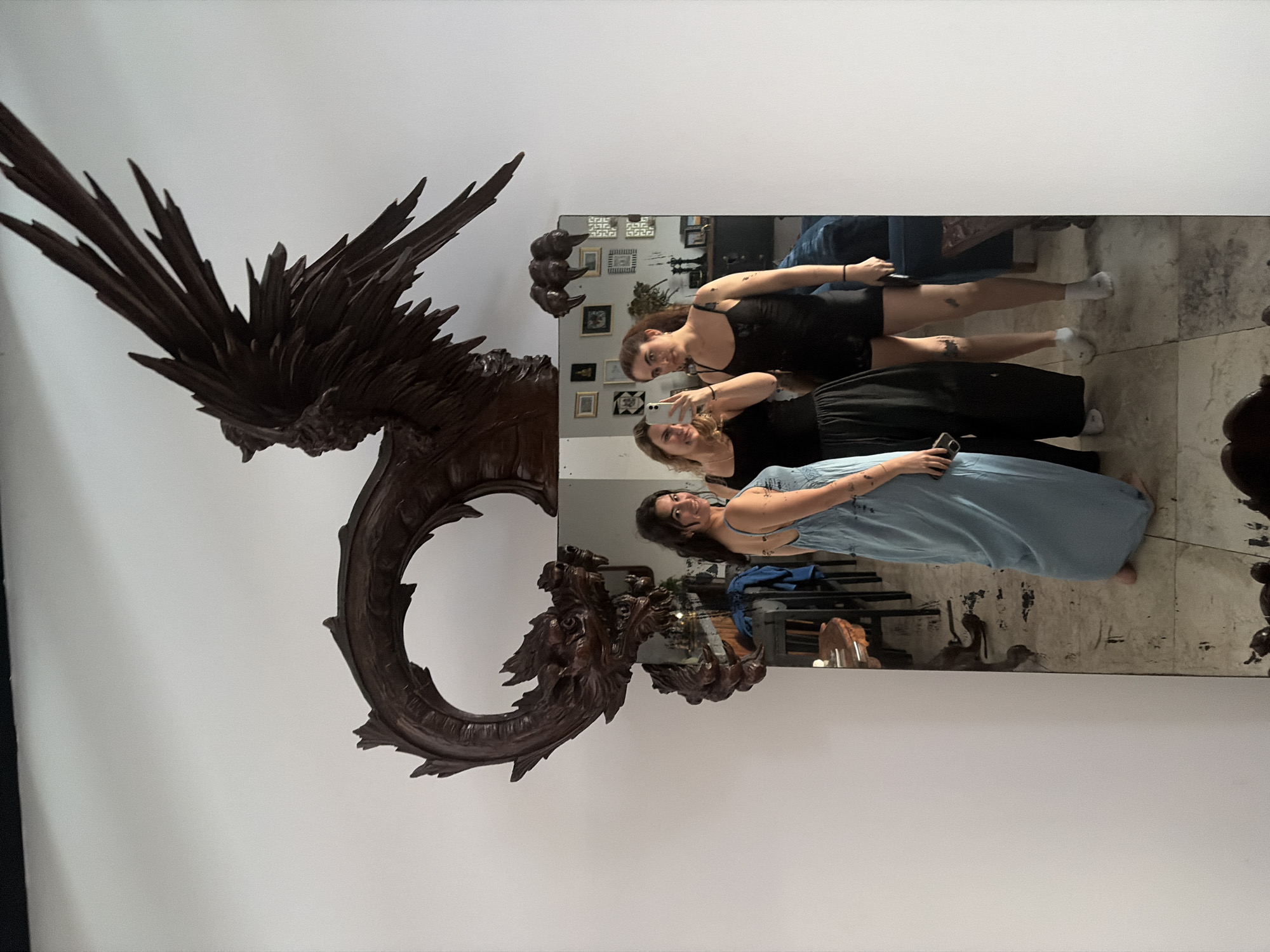 The image shows three women reflected in a large mirror, with an elaborate wooden carving dominating the foreground. The image is taken from a slightly upward angle, capturing the upper bodies of the women and the intricate details of the carving.
In the reflection, three women are visible. One woman, with dark hair, wearing a black sleeveless top and black shorts, is holding a smartphone, seemingly taking a mirror selfie. She has tattoos on her arm and leg. Beside her, another woman with lighter hair, also in a black top, looks towards the phone. The third woman, on the right, wears a light blue dress with straps and has dark hair, looking towards the phone with a subtle smile. Their casual attire suggests a relaxed setting.
The foreground features a magnificent, dark brown wooden carving of a mythical creature, possibly a dragon or a phoenix. Its head, with intricate details, is positioned at the lower right of the carving, and its body curves upwards, culminating in sharply carved wings or feathers that extend towards the top left of the frame. The carving appears to be a substantial piece of art or furniture, perhaps a mirror frame or an decorative element.
The mirror itself has a substantial, dark frame. In the background of the reflection, several framed pictures or artworks are visible on a wall, along with what appears to be part of a shelf or cabinet. The floor visible in the reflection is light-colored, possibly concrete or stone, showing some darker markings.
The activity taking place is a group mirror selfie. The women appear to be in good spirits, casually posing for the photograph. Given the location context of San Bernardino Tlaxcalancingo, Mexico, the large wooden carving might be indicative of local artistic traditions or decor. No discernible text is clearly legible on the phones or in the background reflections.