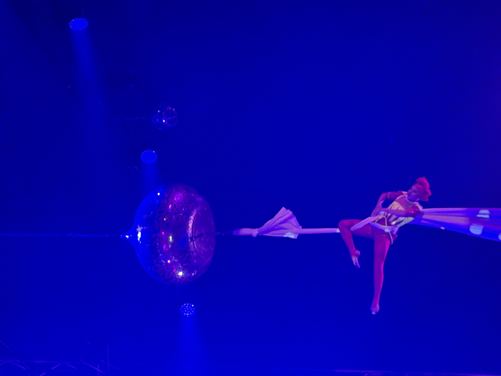 A performer is suspended in mid-air, gripping a long, flowing piece of fabric that is attached to a large, reflective sphere. The sphere is hanging from above and is covered in what appear to be lights or mirrors, giving it a disco ball-like appearance. The performer, wearing a costume with red leggings and a patterned top, is positioned to the right of the sphere, with their body contorted in a dynamic pose. The fabric is draped around them and also attached to a thin horizontal bar that extends from the sphere. The scene is lit with a strong blue light, which casts shadows and highlights the performer and the reflective sphere. There are also smaller, circular light sources visible in the background, also illuminated in blue. The overall impression is of an aerial performance, possibly in a circus or theater setting, taking place in a dimly lit environment with dramatic lighting. No text is visible in the image.