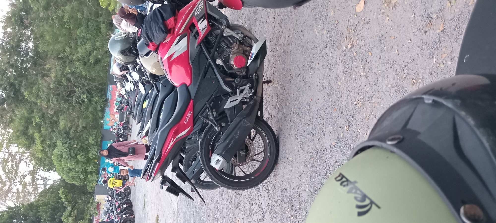 In Mataram, Indonesia, a collection of motorcycles is parked on a gravel surface. In the foreground, a light green helmet with a black trim, featuring a "Vespa" logo, is visible. Behind it, a red and black motorcycle is the central focus, with a silver exhaust pipe and visible engine components. The word "HEART" is legible on the side of the motorcycle. Further back, other motorcycles are parked in rows. A woman wearing a pink headscarf and a pink outfit, holding the hand of a young child in a blue shirt, walks through the area. The background consists of a dense line of trees.