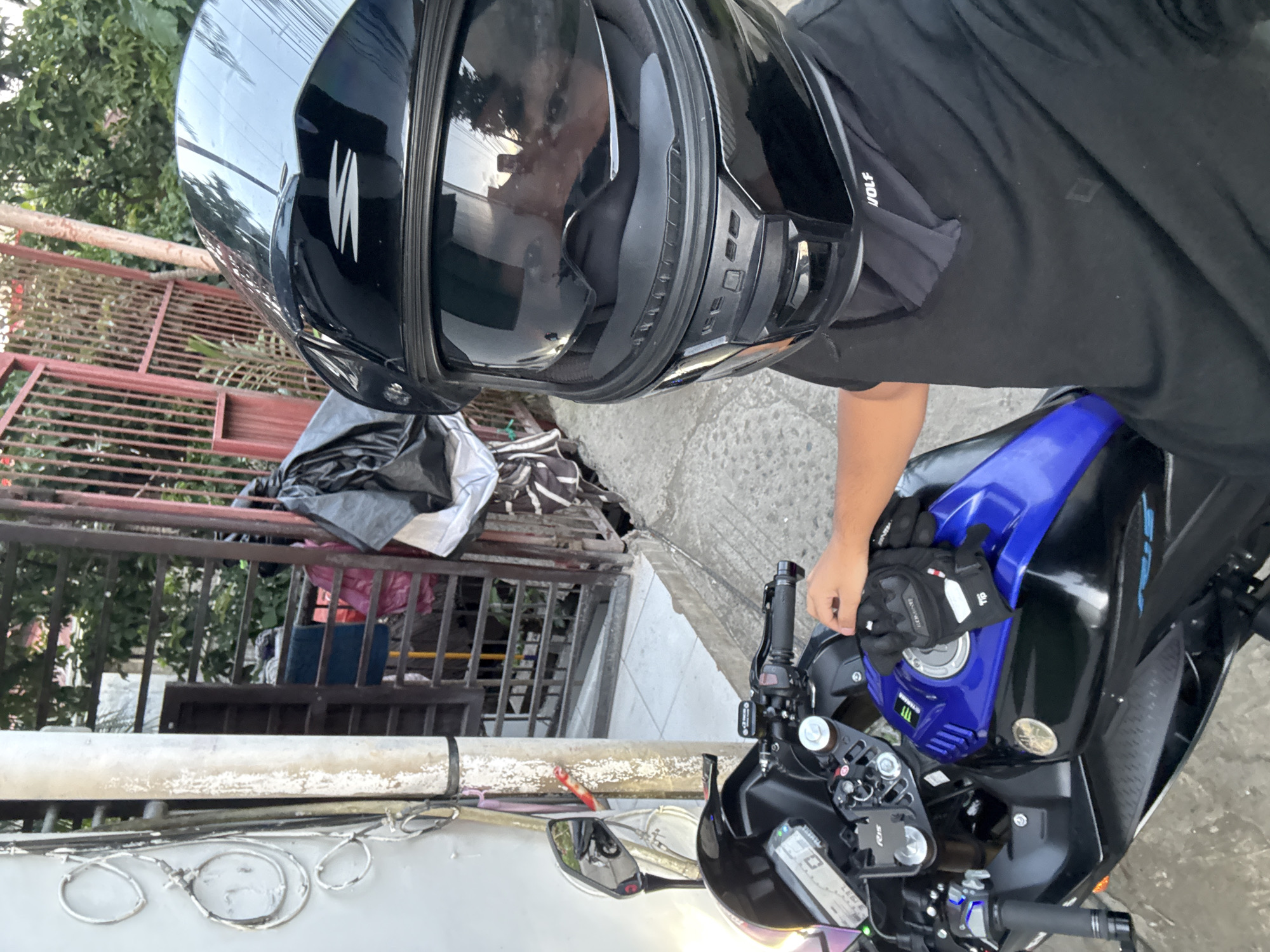 A person wearing a black helmet and black t-shirt is sitting on a blue and black motorcycle. The person's right hand, wearing a black glove, is resting on the motorcycle's fuel tank. In the background, there is a metal gate, a concrete wall, and some trees. The scene appears to be in a residential area, possibly on a street in Davao City, Philippines. The lighting suggests it is daytime.