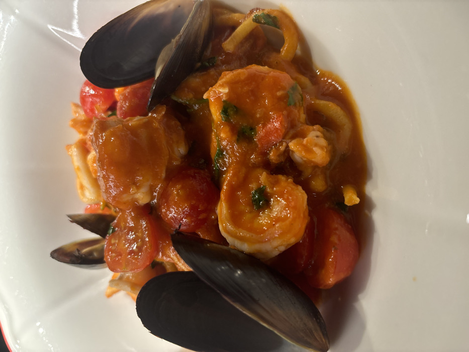 A close-up view depicts a dish of seafood pasta presented in a white, ceramic bowl with subtly fluted edges. The pasta, characterized by thick, yellowish strands, is thoroughly coated in a rich, reddish-orange tomato-based sauce. Visible ingredients include multiple dark-shelled mussels, several cooked pinkish-orange shrimp, and numerous halved red cherry tomatoes. Small fragments of green herbs are interspersed within the sauce. The specific location within Saint Petersburg, Russia, cannot be determined from the visual information provided.