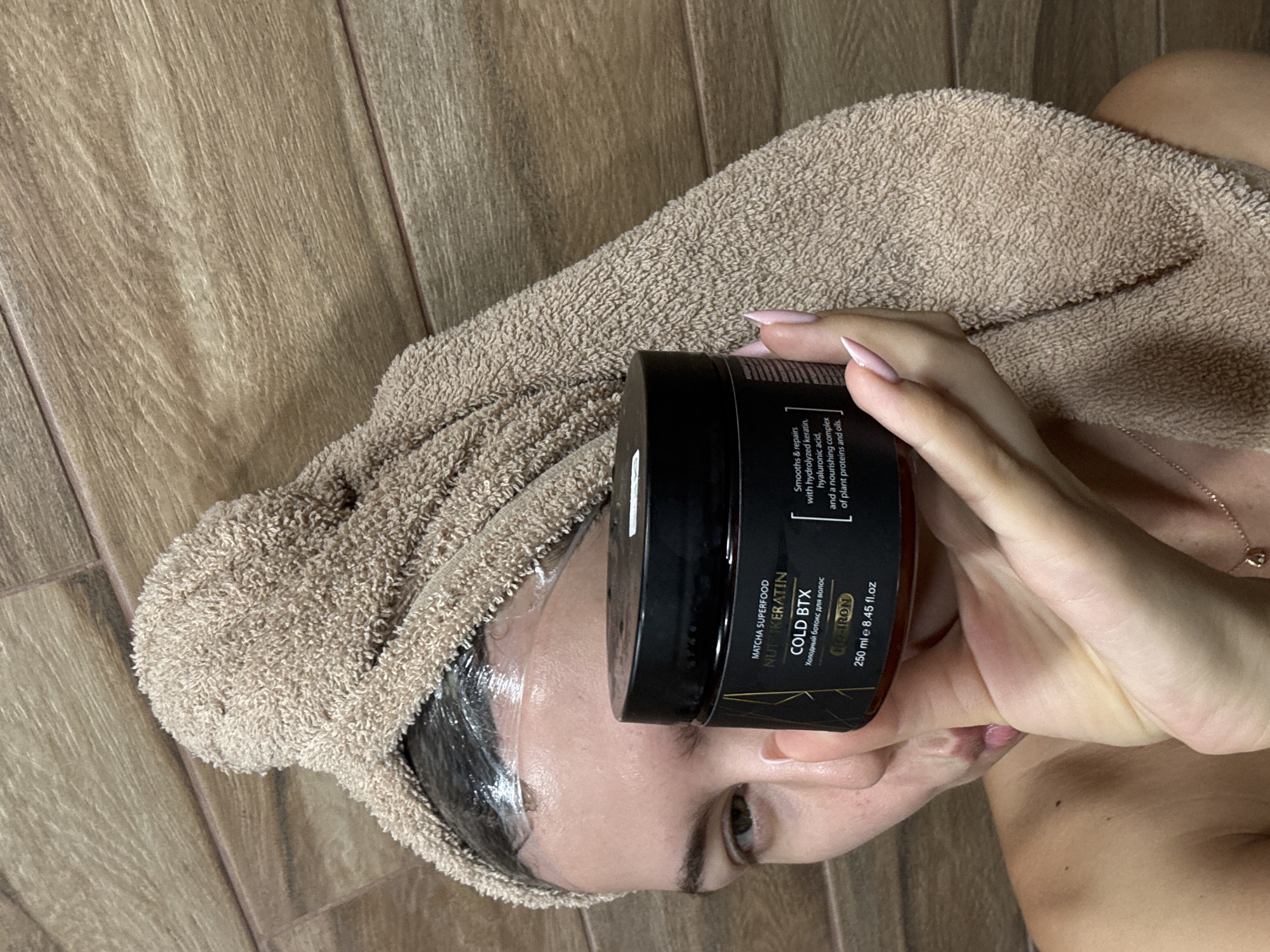 A person with a towel wrapped around their head is holding a jar of hair product. The jar is black with gold lettering and states "MATCHA SUPERFOOD NUTIKERATIN COLD BTX". The background appears to be a wooden wall. The person is looking towards the camera.