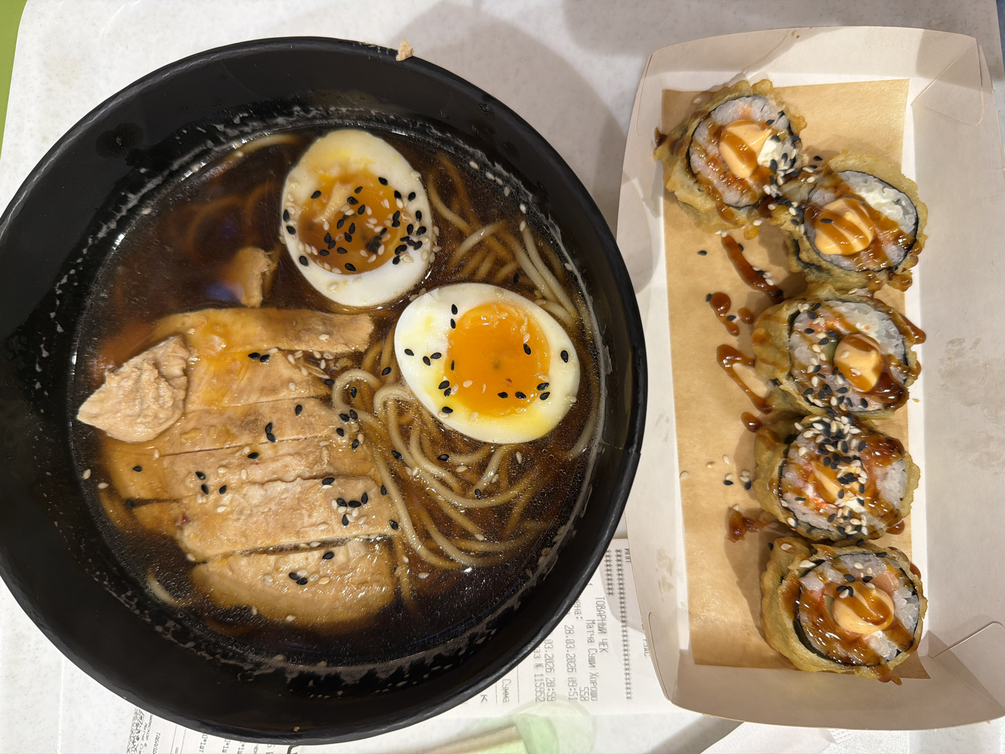 The image shows a meal consisting of ramen and tempura sushi rolls. The ramen is in a black bowl and contains noodles, broth, sliced chicken, and two halved eggs sprinkled with black sesame seeds. The tempura sushi rolls are arranged in a white cardboard container with parchment paper lining, drizzled with sauce, and also sprinkled with black sesame seeds. A receipt from a location named "Matcha Sushi Horosho" dated March 28, 2026, at 09:51, with order number 115952, is partially visible near the sushi container. Based on the OCR, this location is in Russia and likely in the city of Khimki, Russia. The meal is placed on a light-colored surface.