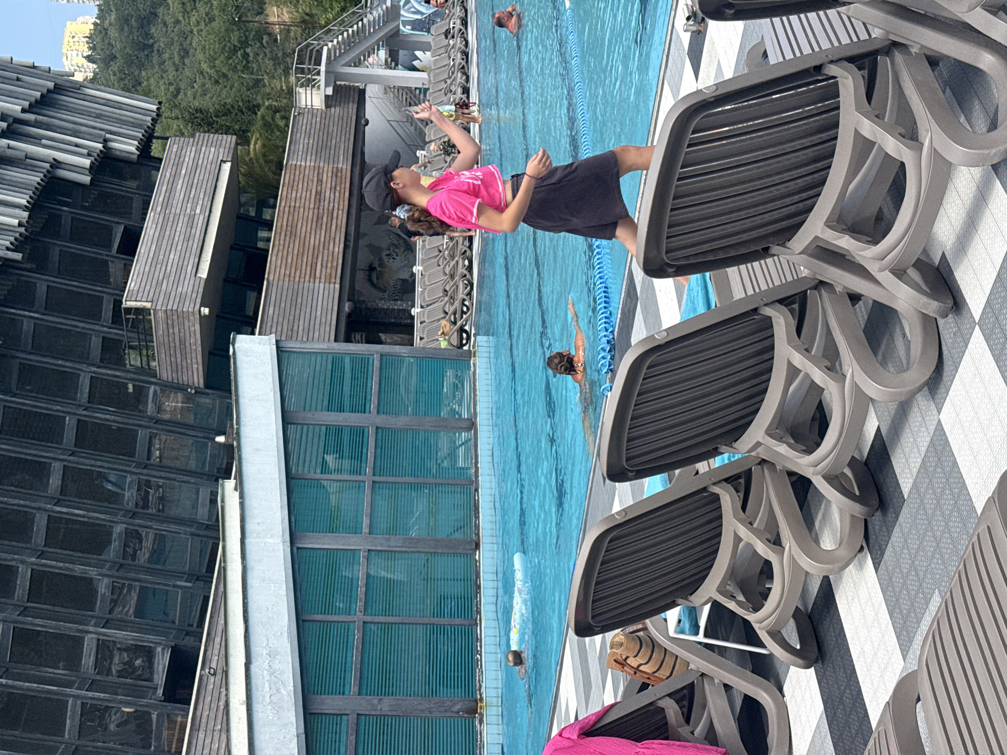 In the city of Massandrovsky village council, Ukraine, a resort swimming pool area is depicted. In the foreground, several gray plastic lounge chairs are arranged on a patterned tile floor. A pink towel is draped over one chair, and a straw bag rests on another. In the middle ground, a blue swimming pool is visible with several people in and around it. A young woman in a pink crop top and dark shorts, wearing a black baseball cap, walks along the edge of the pool, looking to her right. She appears to be holding a smartphone. In the pool, a person is swimming with a flotation device, and another person is partially submerged. Further back, several other lounge chairs are set up, and a large modern building with a distinctive ribbed facade stands behind the pool area, with a lush green hillside in the background. Stairs leading up the hillside are also visible.