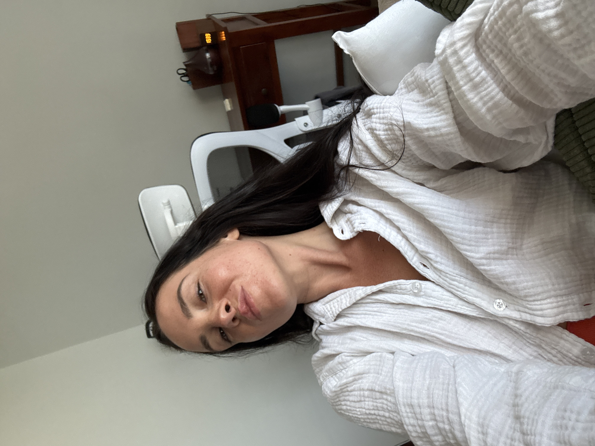 A woman is taking a selfie in a room that appears to be a home office or bedroom. She is sitting, wearing a white gauzy button-up shirt, and her hair is long and dark. In the background, there is a desk with a wooden top, a chair with a white frame and black accents, and a digital clock displaying 19:39. A pillow is visible to her right. The environment suggests an indoor setting.