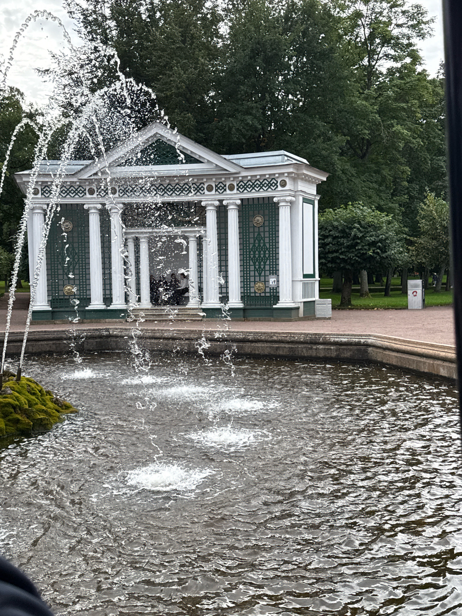 The image shows a fountain in the city of Peterhof, Russia. In the foreground, there is a fountain with water splashing. In the background, there is a white and green structure with columns. There are trees and grass behind the structure.