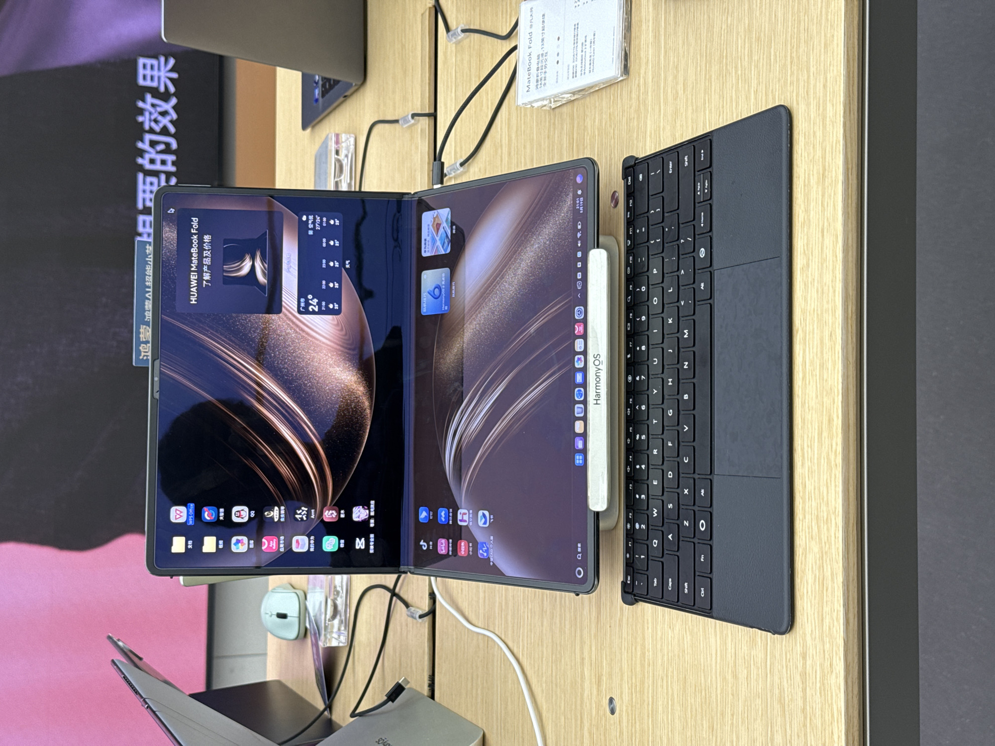 The image shows a Huawei MateBook Fold on display, with a keyboard attached. The screen displays Chinese characters and icons. A stylus labeled "HarmonyOS" is attached to the side of the device. The MateBook Fold is placed on a wooden surface. In the background, a sign with more Chinese characters is visible, alongside other devices on display. The location appears to be a store in Yuexiu District, China.