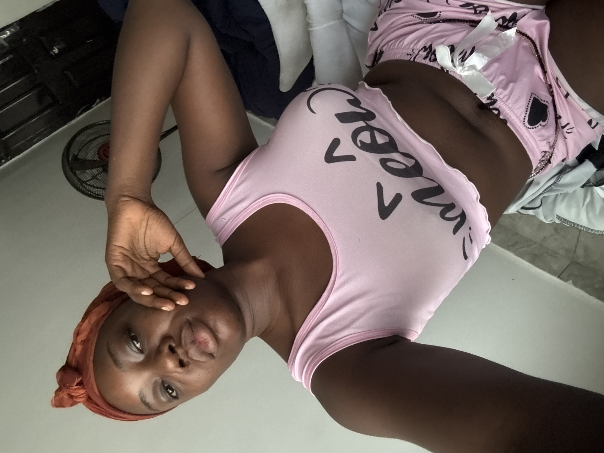 A woman is reclining on a bed in an indoor setting. She is wearing a light pink tank top with a cartoonish design, paired with matching shorts. The room includes a fan and various items like blankets and clothing are scattered in the background. The location is Iddo Tudunwada, Nigeria.