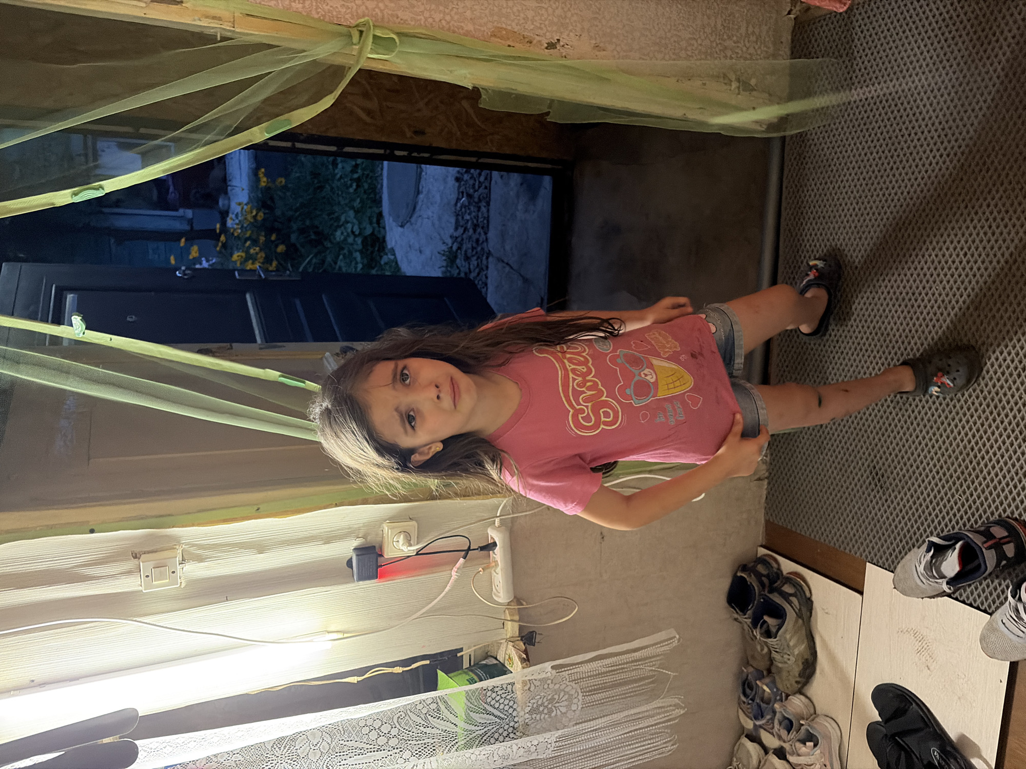 A young girl with long, dark hair and a tearful expression stands in a doorway, wearing a pink t-shirt and denim shorts. Her right leg has visible scrapes. She is inside what appears to be a modest dwelling, possibly a rural home or dacha, in Tambov, Russia. The interior is dimly lit, with a string of lights and electrical outlets visible on the wall to the left. Shoes are scattered on the floor and on white mats near the entrance. A green mosquito net hangs across the doorway, partially obscuring the view of an outdoor area with a dark door and some greenery beyond. The overall impression is one of a domestic, somewhat unkempt environment.