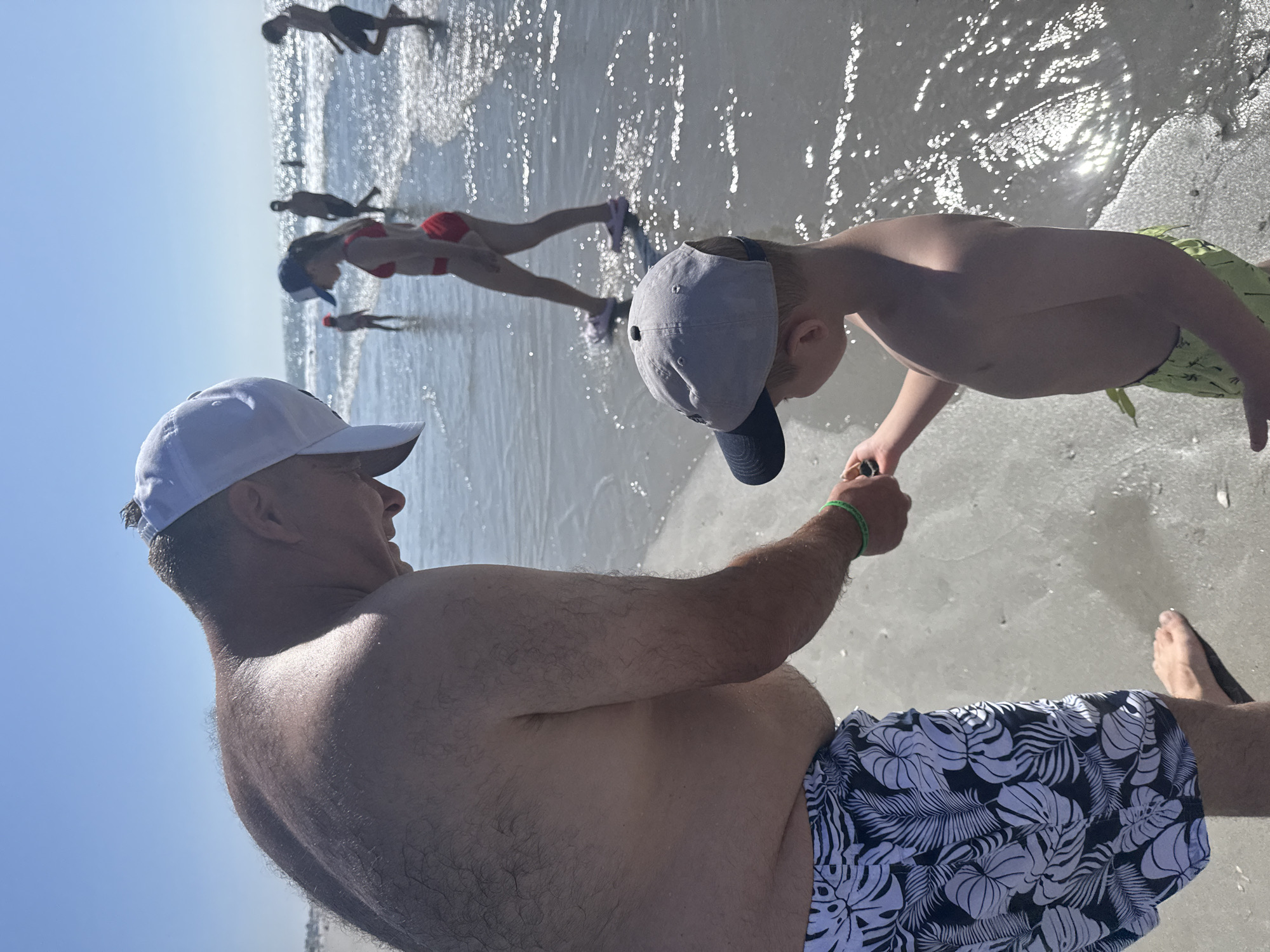 In this image, a man wearing a white cap and patterned swim trunks is in the foreground, facing to the right. He appears to be interacting with a young boy who is also wearing a cap and swim trunks, and is positioned to the man's right. The background reveals a sunny beach scene with several other people in the shallow water. Some individuals are walking or standing, while others appear to be lying down in the water. The water is clear and reflects the bright sunlight, creating a shimmering effect. The sky is a clear blue, indicating fair weather. The overall atmosphere is one of a relaxed day at the beach, with people enjoying the water and the sunshine. The image is oriented vertically, as if taken from a low angle.