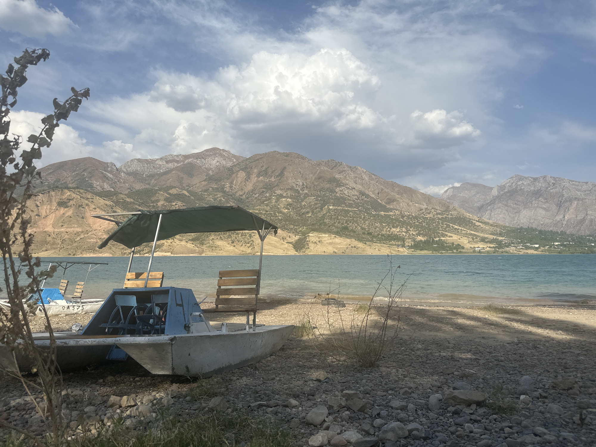 A broad, still shot captures a serene scene at a lakefront, likely within Yusuphana, Uzbekistan. In the foreground, a weathered, light-colored pedal boat rests on a stony shore, its blue pedal mechanism and wooden seats visible under a green canopy. The boat has a distinctive catamaran hull. To the left of this boat, another similar pedal boat with a blue seat and a lighter canopy is partially visible, also beached. The lake's surface is calm, reflecting the blue sky dotted with dramatic white clouds. Beyond the lake, the distant shore is lined with hills and then majestically rises into rugged, brown mountains under a partly cloudy sky. A few sparse, dry plants grow on the rocky shoreline, casting faint shadows on the sand. The overall impression is one of peaceful solitude and natural beauty.