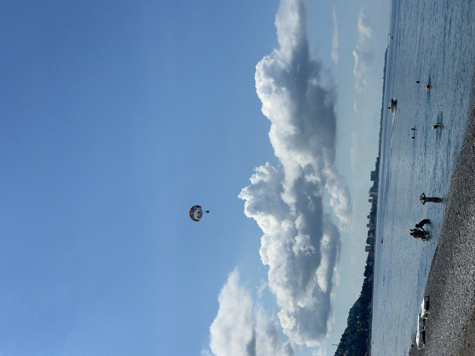A wide-angle, vertical, eye-level shot captures a clear blue sky over a calm sea and a pebbled beach. In the sky, a parasail with a colorful canopy is being towed by an unseen boat, carrying two individuals. Puffy white clouds are scattered across the sky, with a larger formation of clouds nearer to the horizon.
The sea is a tranquil blue, with a gentle shoreline. Several people are visible in the shallow water near the beach, some wading and others swimming. One person stands in the water with their arms raised, while another group appears to be gathered together. In the distance, a motorboat is chugging across the water, leaving a small wake.
Along the distant coastline, a city skyline with numerous buildings is visible, suggesting an urban area. A green, tree-covered hill slopes down towards the city on the left side of the image. The beach in the foreground is made up of small, gray pebbles, with a few empty white lounge chairs placed on it. The overall scene depicts a leisurely summer day at the beach, with recreational activities taking place on both the water and in the air. The location is Abaata, Abkhazia.