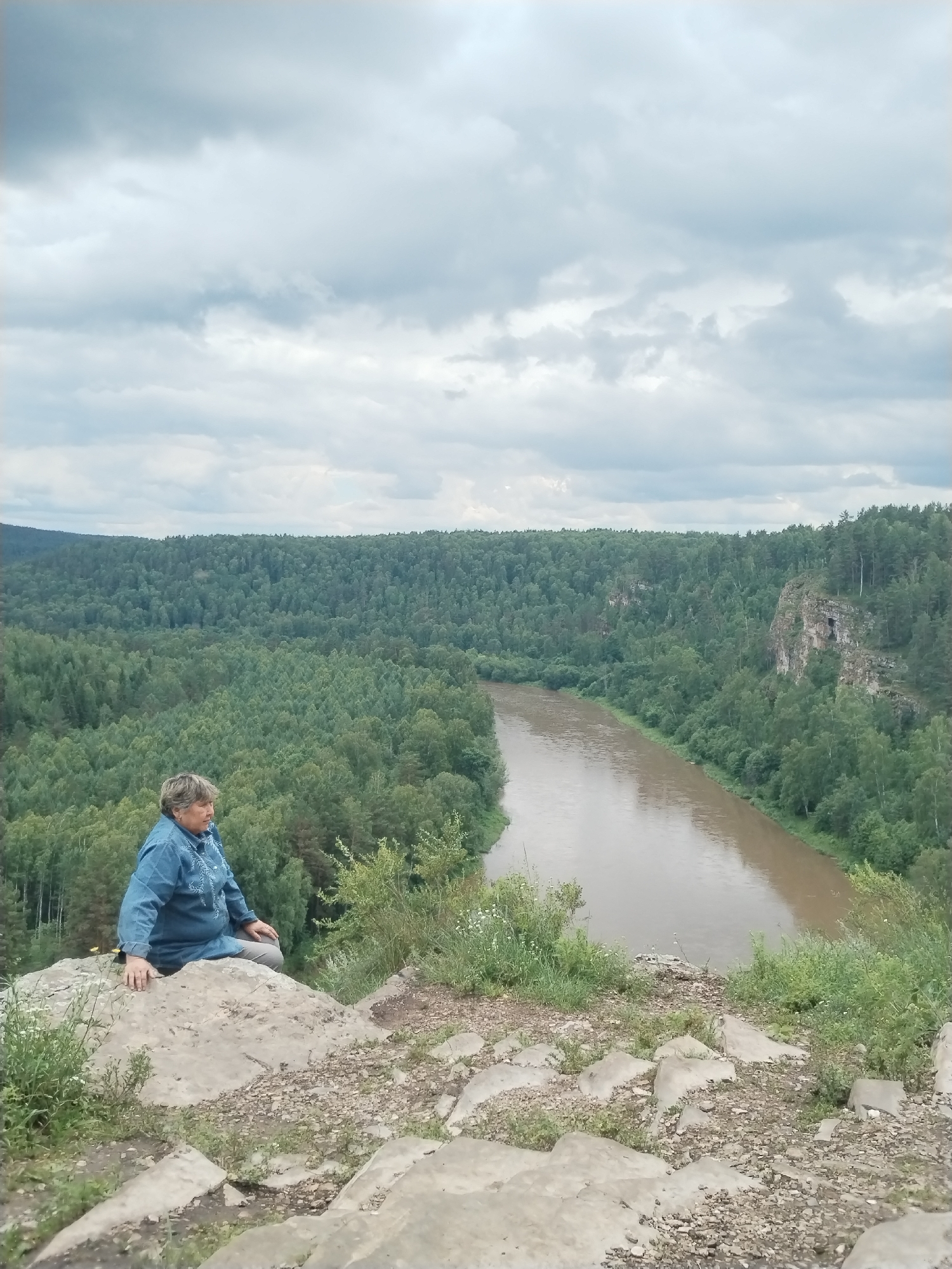 A woman is sitting on a rocky outcrop overlooking a wide, muddy river that winds through a dense forest. The sky is overcast with dramatic, grey clouds. The woman is wearing a denim jacket and grey pants. Her hands are resting on the rock in front of her, and she is looking out at the view. The foreground is rocky and sparse with some green vegetation, while the background is dominated by the thick green canopy of the forest and the river. The location appears to be a natural scenic overlook, likely in a rural or wilderness area, such as the vicinity of the city of Idrisovo, Russia, known for its natural landscapes.