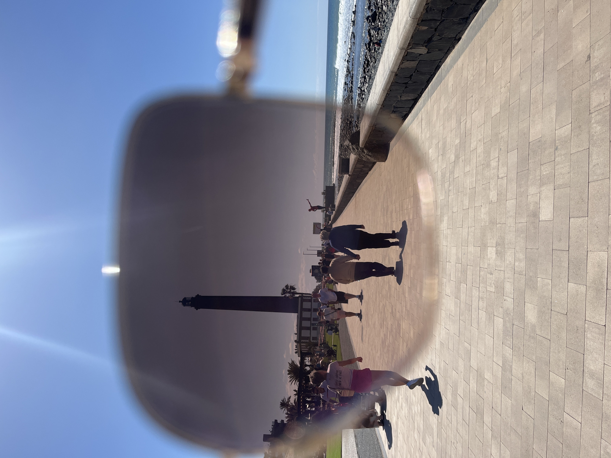 The image captures a scene in San Bartolomé de Tirajana, Spain, focusing on a paved walkway along the coast. People are walking and exercising in the sunlight. The Maspalomas Lighthouse is seen through the lens of glasses, adding a distorted layer to the composition. Palm trees, a low stone wall, and the ocean with a rocky shore are visible in the background.