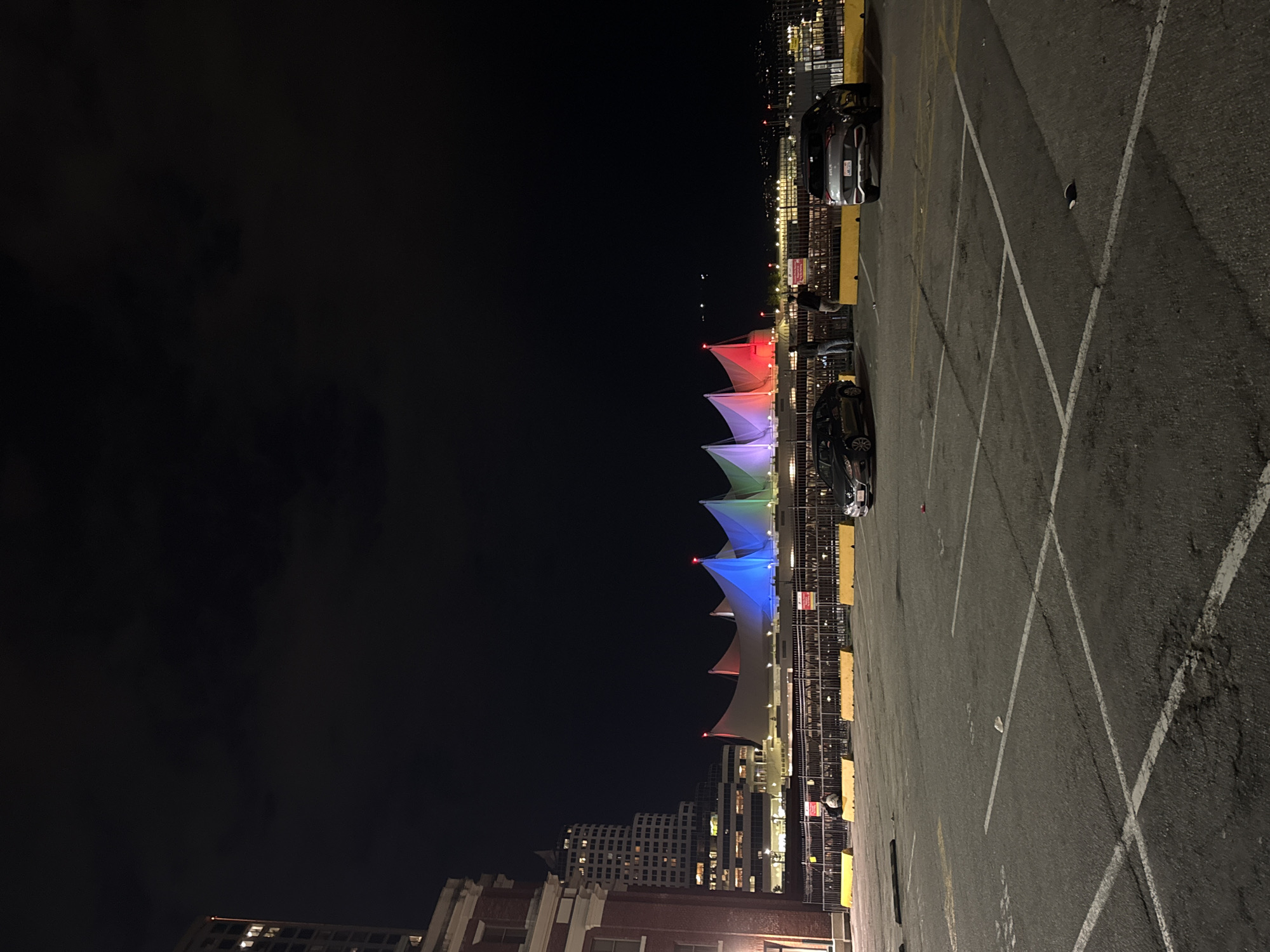 The scene is an outdoor night shot, likely from a rooftop or elevated parking structure in Vancouver, Canada. The focal point is Canada Place, identified by its distinctive white sail-like structures illuminated with red, green, and blue lights. The lighting pattern suggests it's potentially for a special occasion or event.
In the foreground is a paved surface, potentially a parking area. It is marked with white parking lines and shows signs of wear and tear. Two cars are parked along the edge of the parking area. There are also yellow barriers or curbs separating the parking area from the edge. Several people are standing along the edge, appearing to be observing Canada Place.
In the background, a skyline with buildings of varying heights and illumination levels is visible. Some of the buildings are modern high-rises, and others are older brick structures. The sky is dark, indicative of nighttime. There is some cloud cover.