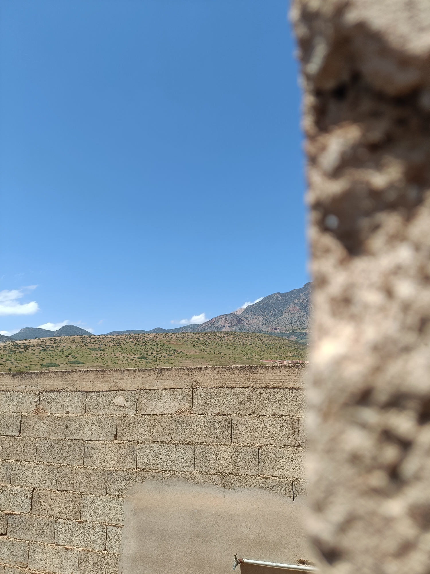 The image, likely taken in Ouaouizert, Morocco, features a cinder block wall with a partially plastered area on the lower section, as well as a metal pipe and faucet. Beyond the wall, there is a view of rolling, sparsely vegetated hills and a distant mountain range under a clear blue sky with a few scattered clouds. A blurry, textured object, possibly a stone structure, is in the foreground on the right side of the image, partially obscuring the view. There are no visible people or interactions.