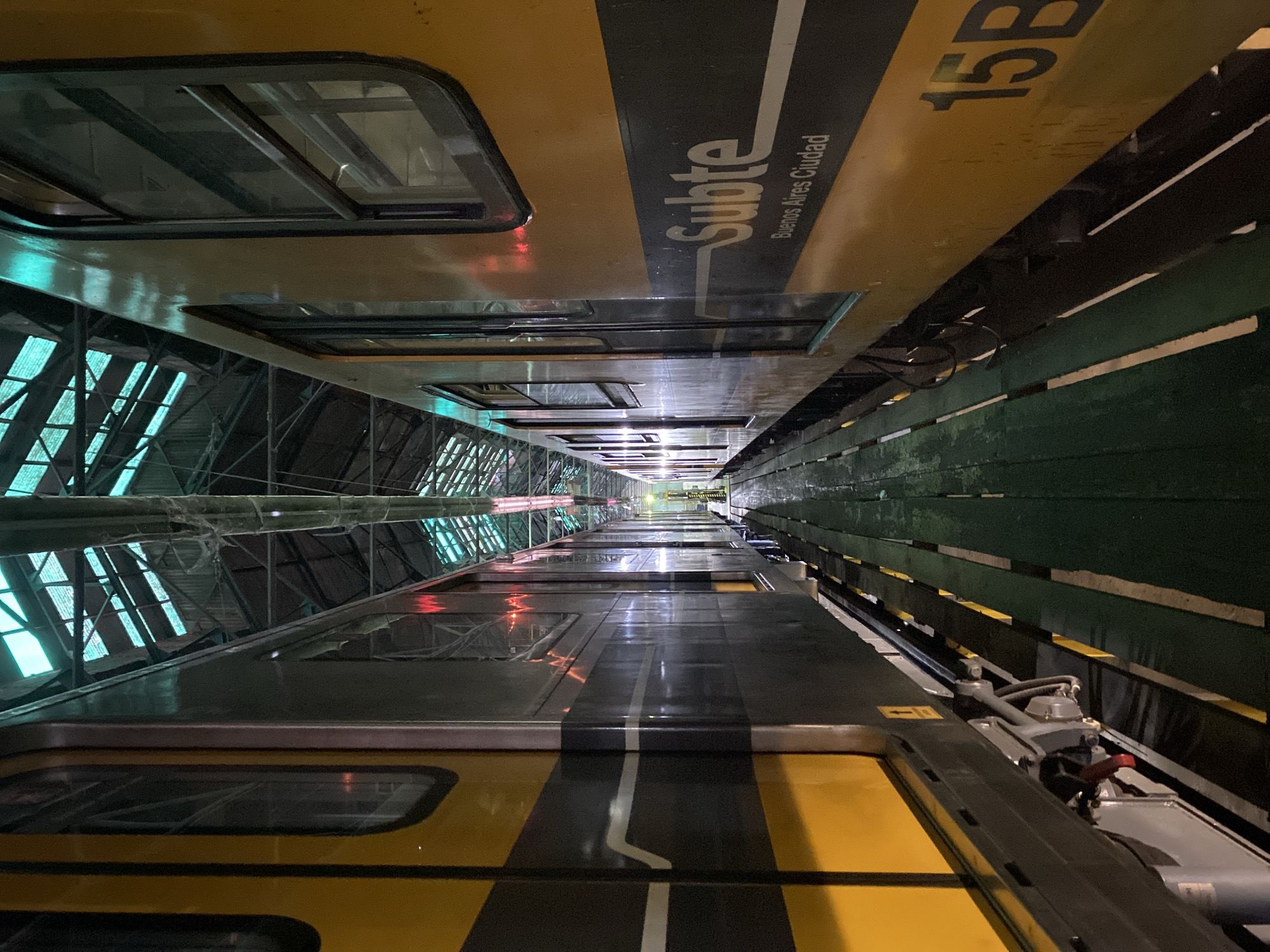 This image depicts two Buenos Aires Subte (subway) trains parked parallel within a maintenance or storage facility in Buenos Aires, Argentina. A narrow, dark green wooden slatted walkway separates the trains, revealing tracks and undercarriage components beneath.
The train on the left is predominantly dark gray with a prominent horizontal yellow stripe and a thinner white stripe along its side. Its windows are dark and reflective.
The train on the right features a primary yellow livery with a horizontal dark gray stripe bearing the white "Subte" logo and "Buenos Aires Ciudad" text. The numeric designation "15B" is visible in dark characters on the lower yellow section of this train. Its windows are also dark.
The high-ceilinged facility features a complex overhead structure of metal trusses and translucent or corrugated panels, through which a diffused greenish-blue light penetrates. Artificial lighting sources, some emitting a reddish glow reflected on the train surfaces, illuminate the interior. The environment is industrial, with exposed tracks and dark, potentially wet, surfaces beneath the trains. No individuals, actions, or interactions are visible.