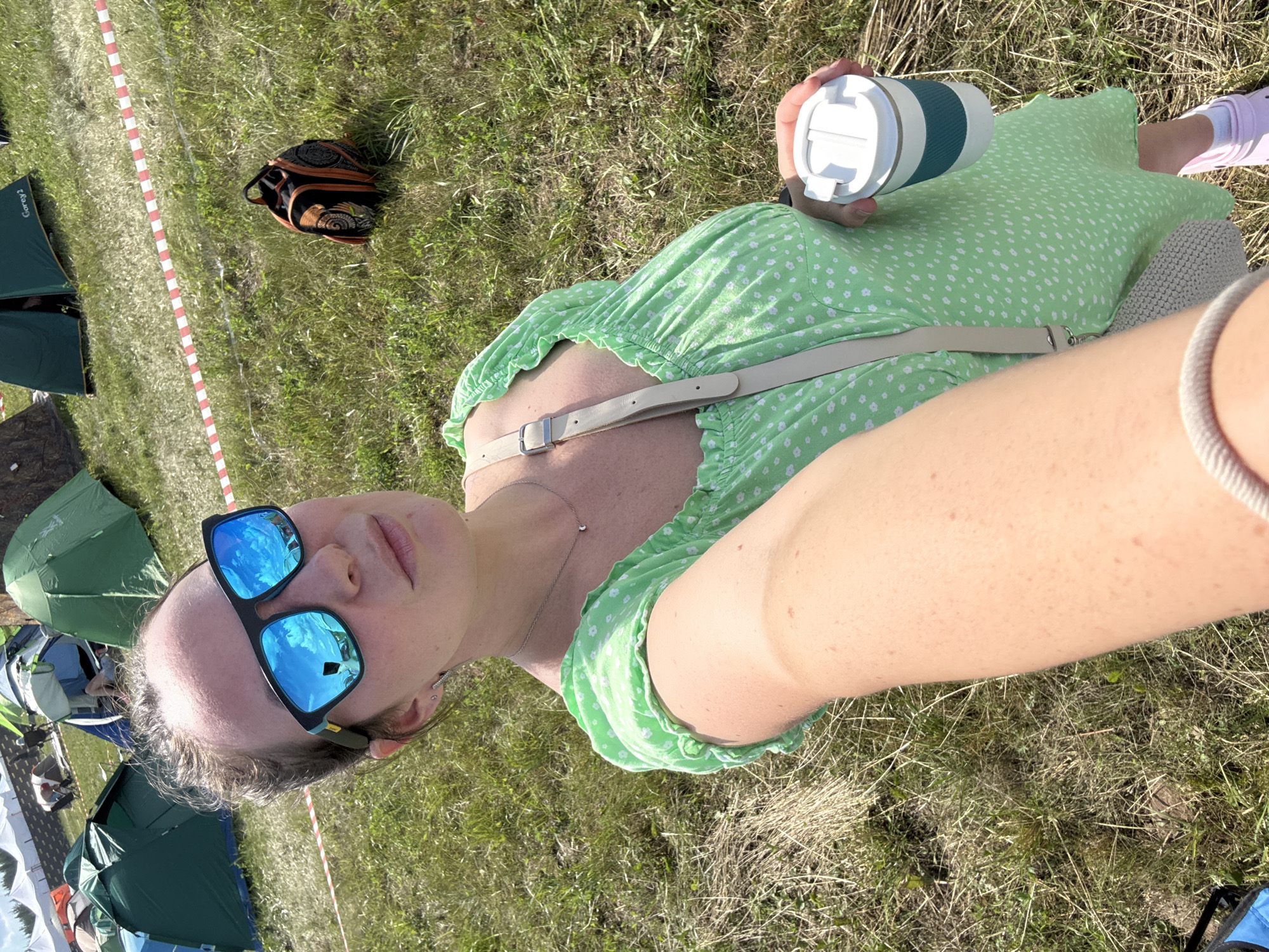 In a grassy field with tents set up, a woman takes a selfie. She is wearing a light green dress with white polka dots, sunglasses, and a light-colored crossbody bag. In her right hand, she holds a white travel mug with a teal band. Her left arm is extended towards the camera, and her right arm is holding the mug. The background shows several tents of various colors, some with people inside or near them. A red and white striped caution tape runs horizontally across the middle ground. The ground is covered in dry grass and some patches of green. The sky is visible at the top of the frame and appears to be blue with some white clouds. This scene appears to be at an outdoor event or campsite.