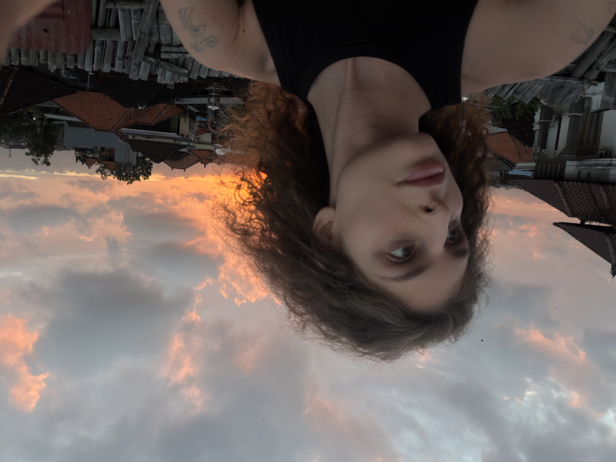 The image, taken in North Kuta, Indonesia, features a person looking at the camera against a backdrop of a sunset sky and a village. The person, with curly brown hair, is wearing a black tank top. Their face is partially visible, with one eye and part of their mouth showing. The sky is filled with clouds illuminated by the warm hues of the setting sun, creating an orange and pink glow.
Below the sky and behind the person, the scene depicts a village with rooftops of houses. The architecture suggests a tropical location, with tiled roofs and some bamboo structures visible. The overall atmosphere is serene, with the sunset casting a peaceful ambiance. The image appears to be a selfie, taken from a high vantage point, possibly a rooftop. The time of day is clearly sunset, indicated by the colors in the sky. There is no visible text in the image.