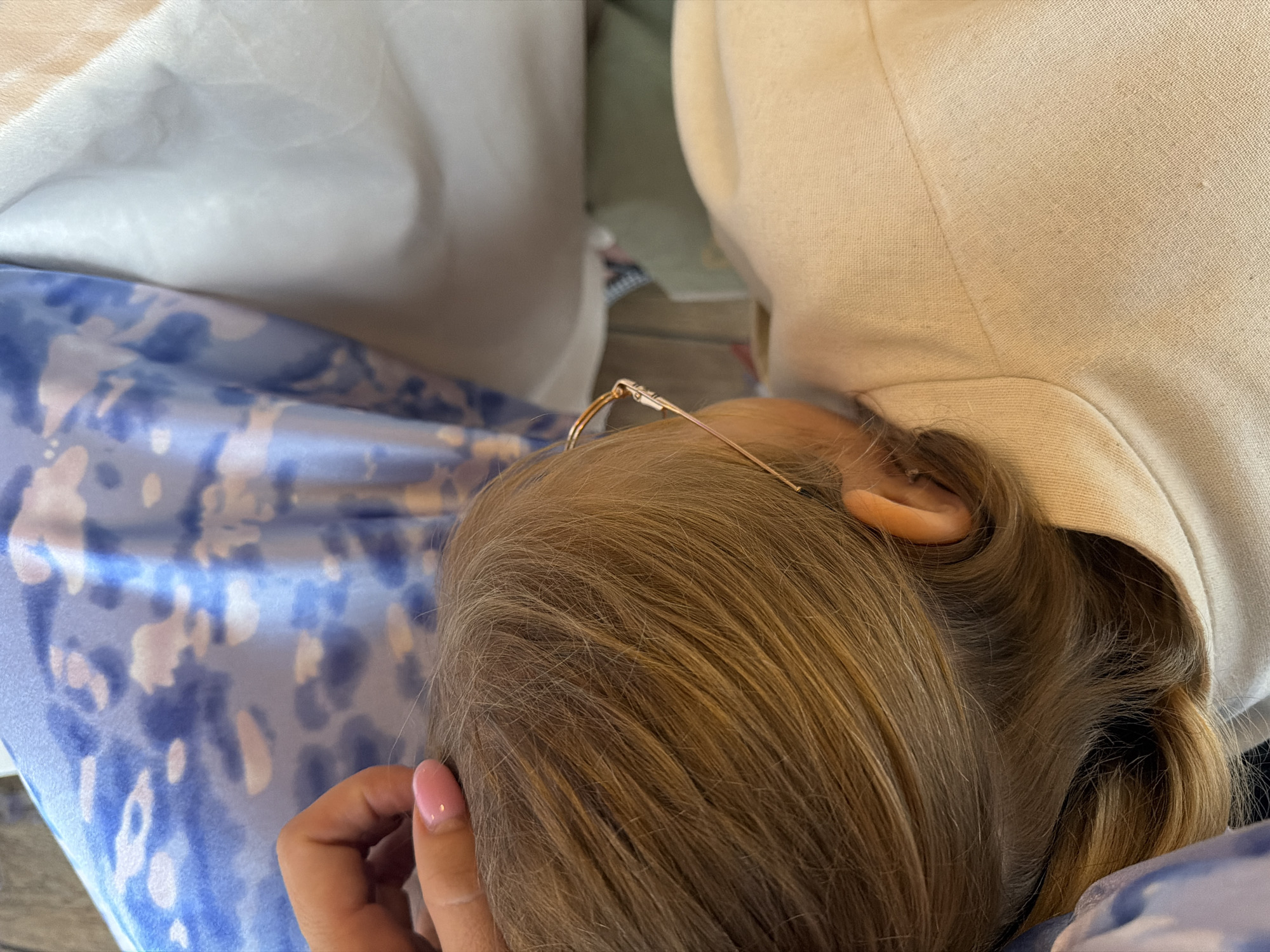 The image shows a close-up view of a person's head from behind, resting on a light-colored garment that appears to be a jacket or sweater. The person has blonde hair and is wearing thin-rimmed glasses that rest on their head. A portion of their ear is visible, as is a hand with a pink-painted fingernail touching their hair. The background is out of focus but suggests a soft, indoor setting with draped fabric in shades of white and blue with a tie-dye pattern. The lighting is soft and diffused, indicating it might be daytime, but no specific time of day can be determined. There is no visible text in the image, and no strong emotional cues are evident, other than perhaps a sense of relaxation. The context provided, Domodedovo, Russia, does not offer any specific visual cues within the image itself.