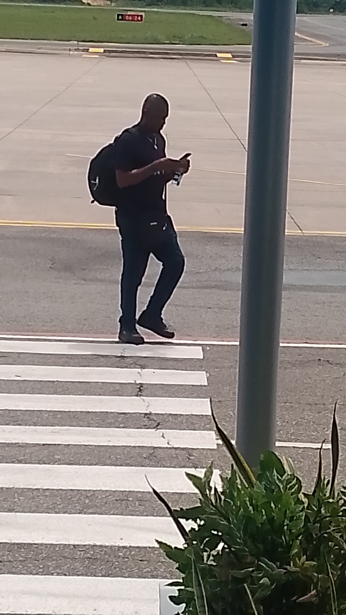 An adult male wearing a black t-shirt, dark pants, and dark shoes is walking across a pedestrian crossing at an airport. He is carrying a black backpack and holding a smartphone in his right hand, looking down at it. The environment appears to be an outdoor tarmac area with a gray concrete surface and white pedestrian crossing lines. A tall gray pole is visible to the right of the frame, and some green foliage is in the foreground on the right. In the background, there is a red sign with white lettering that reads "A 06:24". The scene is lit by daylight.