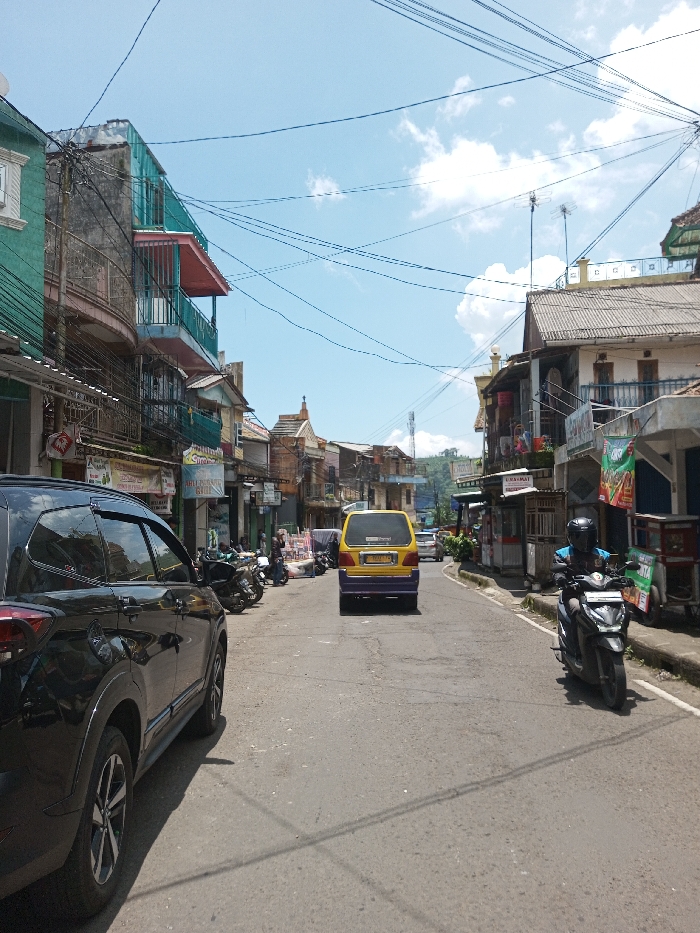 The scene depicts a daytime street view in the city of Bagochen Boolingpang Ueling, Bhutan, under a clear blue sky with scattered white cumulus clouds. Numerous black utility wires crisscross overhead.
The asphalt road, showing signs of wear and patching, extends into the distance, gently ascending before curving out of view. Concrete sidewalks line both sides of the street.
On the left, a black SUV, possibly a Toyota Rush, is parked, facing away from the viewer, with its rear driver's side visible in the foreground. Behind it, several motorcycles are parked on the sidewalk in front of multi-story buildings. These buildings exhibit varied architectural styles, featuring balconies with metal railings, some painted in teal, pink, and faded yellow. Ground-floor storefronts display various signs, including "Susu Murni" and "ABILLY STORE," with dense utility wiring affixed to their facades.
In the center of the road, a yellow and purple van, possibly a public transport vehicle, moves away from the viewer. On the right side of the road, a dark-colored scooter/motorcycle is approaching, operated by an individual wearing a dark helmet and a blue top. The right side of the street is also lined with multi-story buildings, one featuring a dark corrugated roof and a balcony with dark blue railings. Storefronts are present, some with awnings and outdoor displays. A red and white banner is visible on the far right sidewalk, and a partially visible "INDOMARET" sign suggests a convenience store. Antennae are visible on some rooftops. Indistinct figures are visible on sidewalks further down the street.