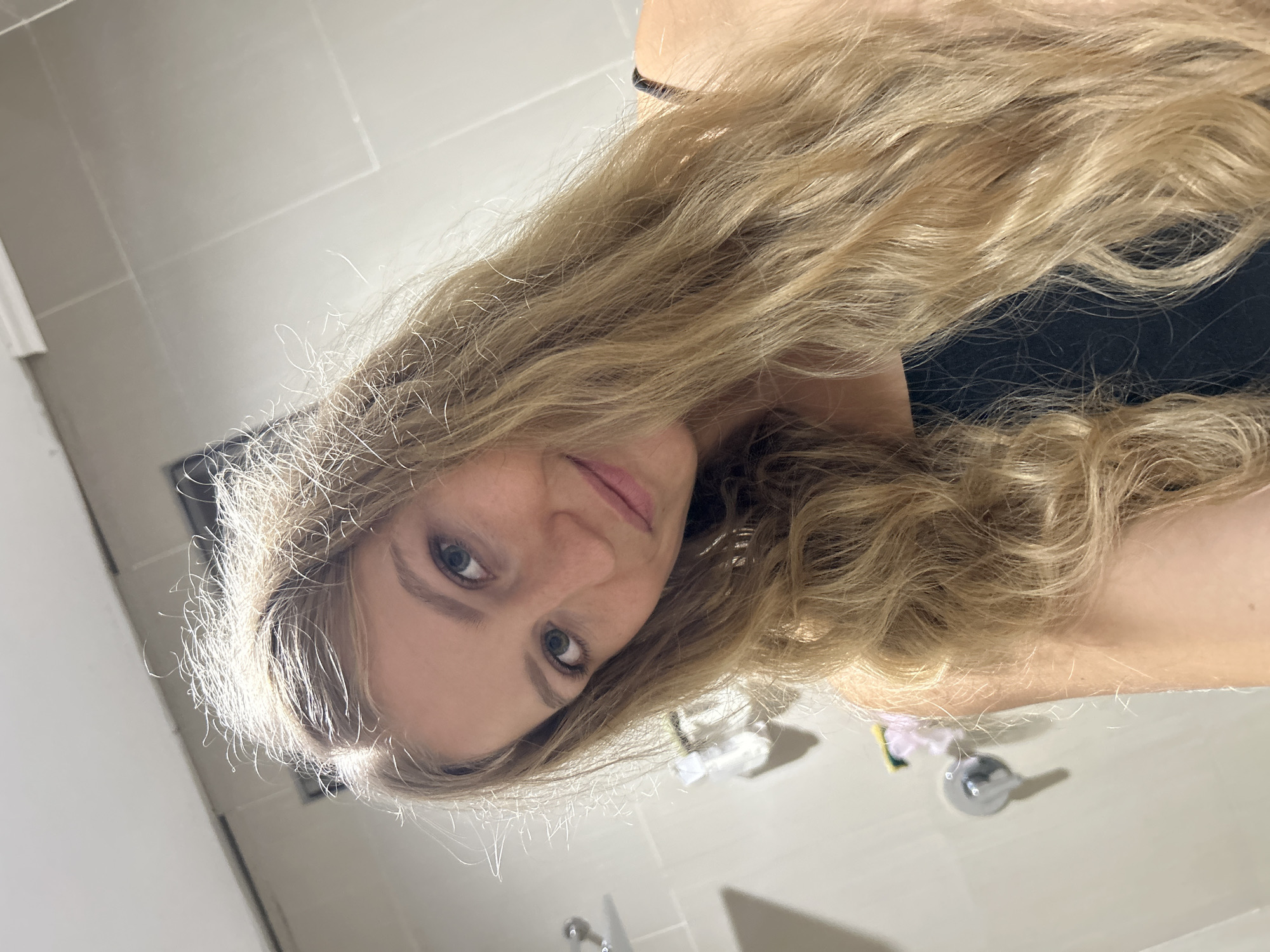 A young adult female is depicted in a rotated, close-up selfie taken indoors, likely in a bathroom in Kuta Selatan, Indonesia. She has long, wavy, golden blonde hair that extends across much of the frame, with individual strands catching the bright light, particularly around her face. Her hair appears to be in good condition with some natural texture and volume. She has light-colored eyes, possibly blue or green, and a natural, pleasant expression with a slight, subtle smile. Her gaze is directed upwards and slightly to the side, looking directly at the camera. She is wearing a dark-colored top, possibly black, which is partially visible beneath her hair and around her shoulder.
The background consists of light-toned, possibly off-white or light gray, tiled walls, indicative of an indoor setting like a bathroom. Reflections of light are visible on these tiled surfaces, suggesting a bright light source overhead or nearby. In the lower right corner of the original image, a silver fixture, possibly a faucet or a knob, is visible on a flat surface, further supporting the bathroom setting. Some indistinct small objects, possibly toiletries, can also be seen in the background behind her head. The overall lighting is bright and even, illuminating her features and hair. There is no visible text in the image.