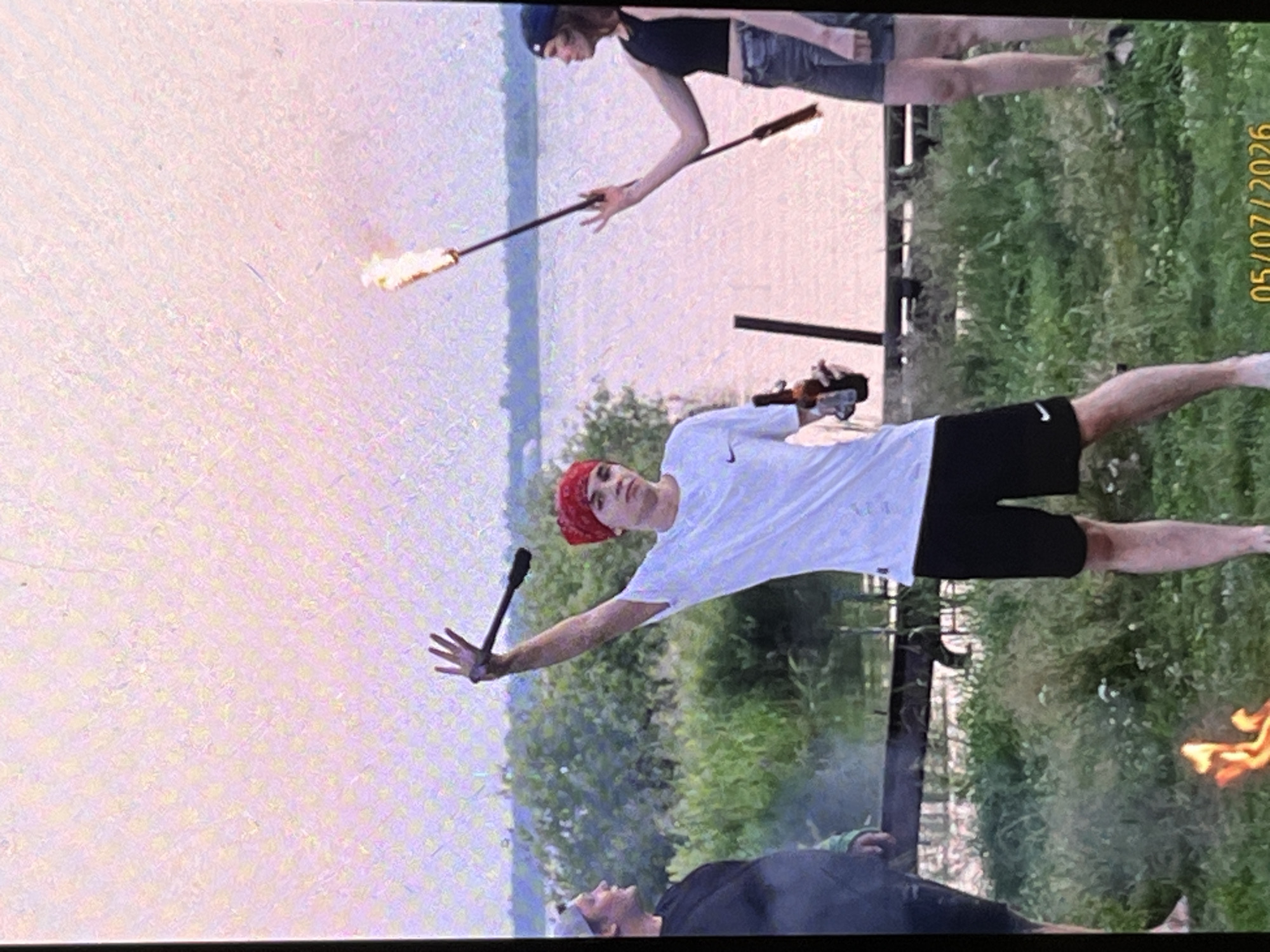 A group of people appear to be gathered outdoors by a body of water, possibly a lake or river, at dusk or dawn. A young man in the foreground is wearing a white t-shirt, black shorts, and a red bandana on his head. He is holding a flaming torch in his left hand and a bottle of beverage in his right hand. To his left, another person wearing a black shirt and a gray hat is visible. To his right, a young woman is holding a flaming torch. A small fire is burning on the ground in front of the man. The background consists of trees and a body of water with a soft pinkish hue, suggesting either sunrise or sunset. The image has a grainy quality and a timestamp in the bottom right corner reads "05/07/2026". The overall atmosphere appears to be casual and celebratory. The location is identified as the city of Tyumen, Russia.