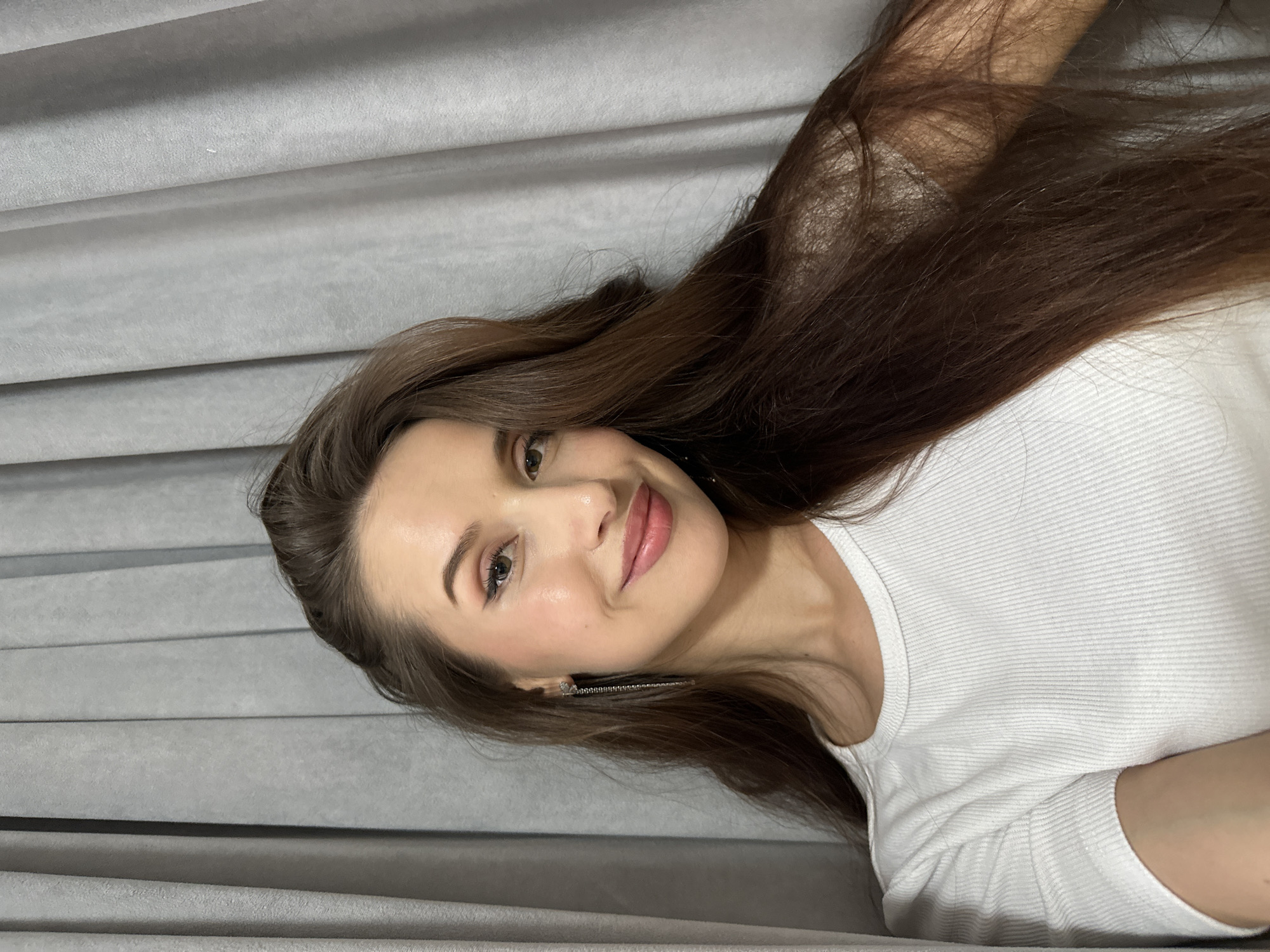 A woman with long brown hair and light skin is lying down, facing the camera. She is wearing a white shirt and long, dangling earrings. The background appears to be a gray, textured fabric, possibly curtains or upholstery. There is no evidence to suggest a location in Yekaterinburg, Russia.