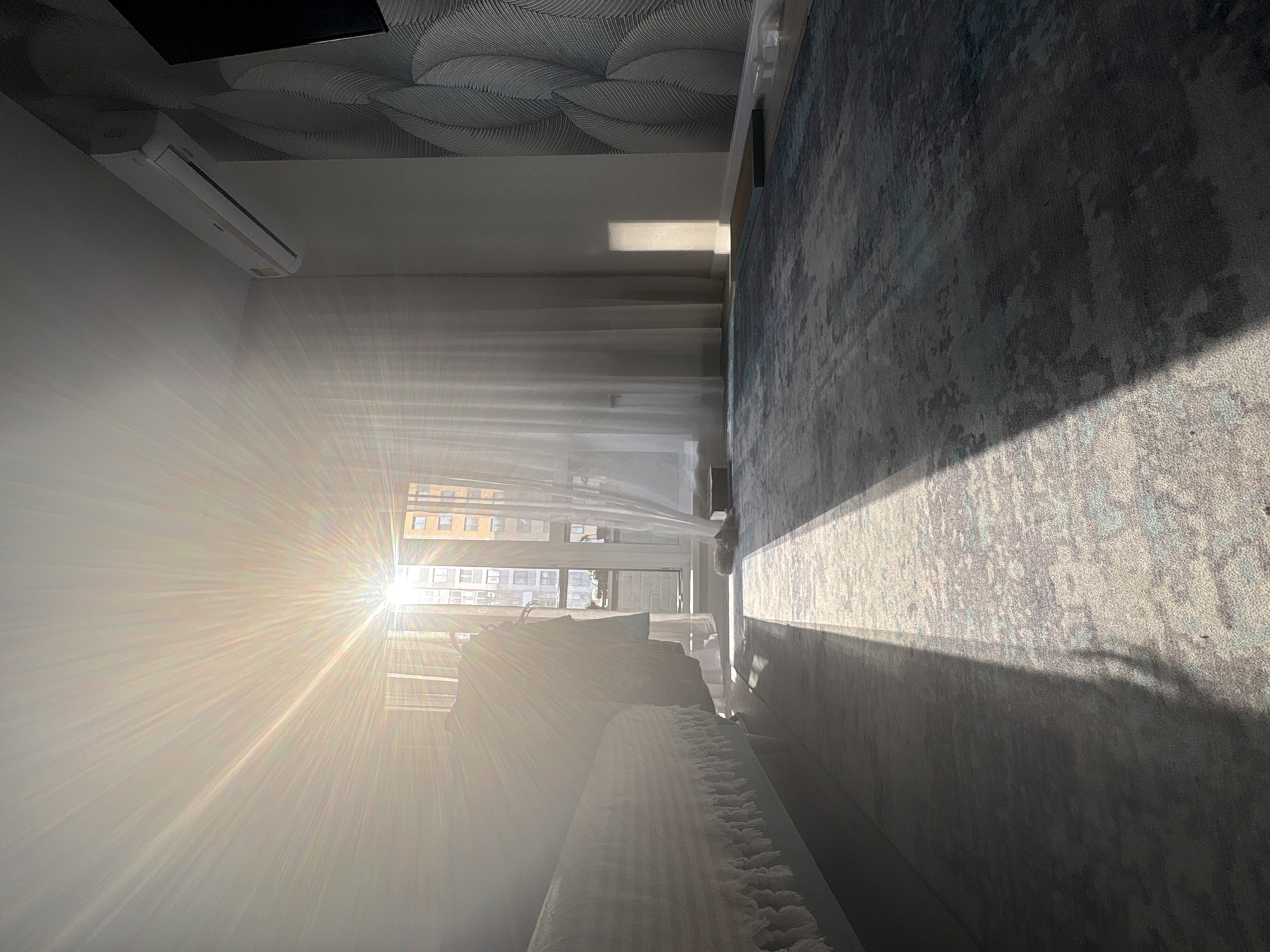 The image shows the interior of a room, heavily illuminated by sunlight streaming in through a window. Outside the window, buildings are visible, suggesting an urban environment. The room features a bed with a patterned blanket, a patterned ceiling, a rug, and a wall-mounted air conditioning unit. The strong sunlight creates a bright, overexposed effect within the room. The picture was likely taken in the city of Izhevsk, Russia.