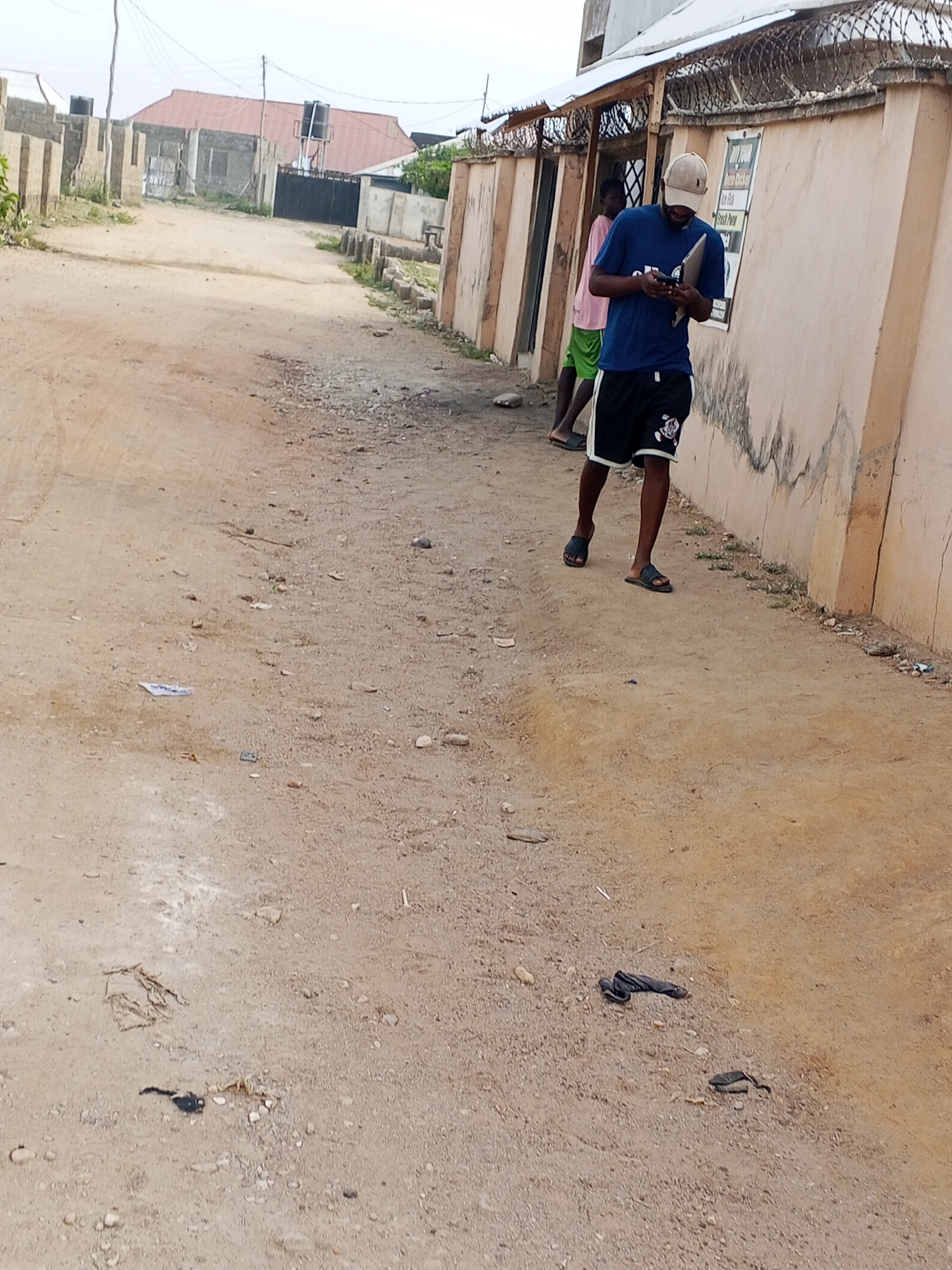 Location: Otokiti, Nigeria. A dirt road runs between buildings. A man wearing a blue shirt, black shorts, and a light-colored baseball cap is standing and looking at a mobile device near a light-colored wall. Another person in a pink shirt and green shorts stands behind him. Debris, including dark cloth-like material, is scattered on the ground. A building with a red roof is visible in the distance.