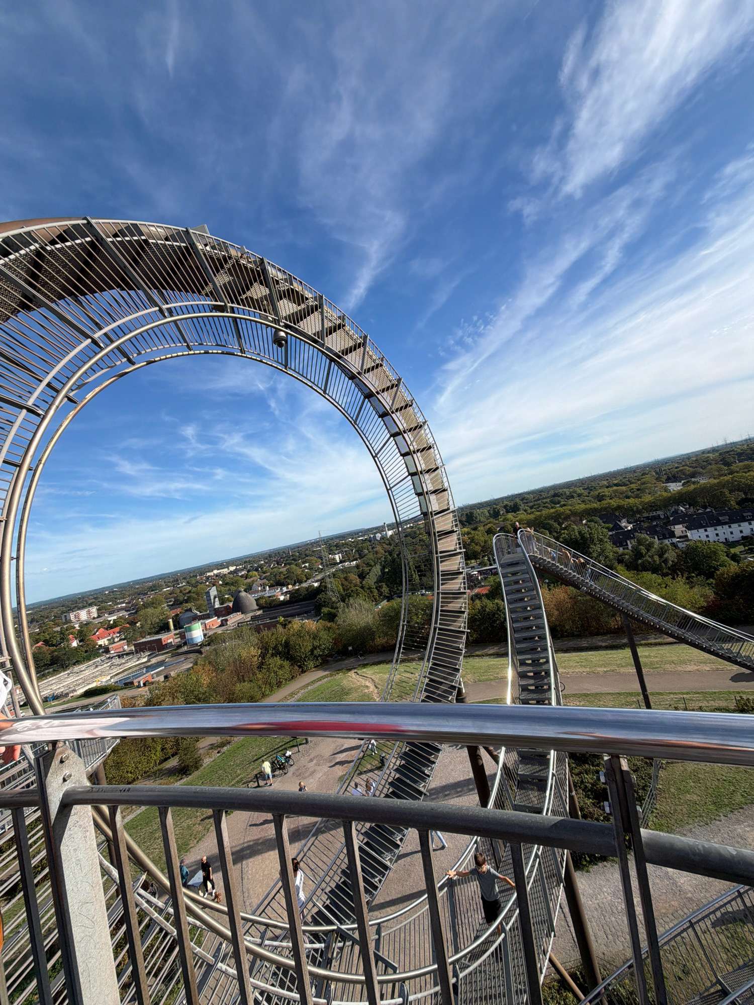 The image shows a panoramic view from the top of the "Tiger & Turtle - Magic Mountain" sculpture in Duisburg, Germany. The sculpture, a large metal structure resembling a roller coaster, dominates the foreground with its spiraling ramps and staircases. The sky above is blue with wispy white clouds. Below, the city of Duisburg unfolds, a mix of residential buildings, industrial structures, and green spaces, including trees and a body of water. Several people are visible on the sculpture, some walking up or down the stairs, while others are observing the surroundings. The scene appears to be set on a clear day, likely during daylight hours, with the sun casting shadows on the metalwork. The overall impression is one of architectural marvel set against an urban and natural landscape.