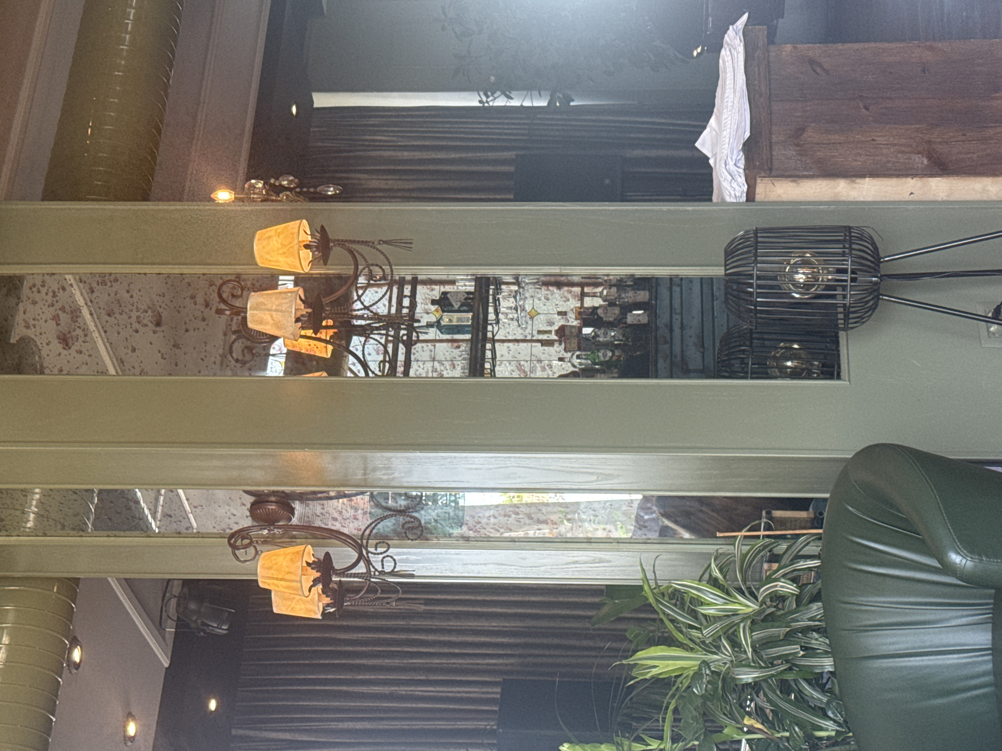 The image depicts an interior scene in a dimly lit establishment, possibly a bar or restaurant, located in Sabail Raion, Azerbaijan. A large, floor-to-ceiling mirrored column dominates the center of the frame, reflecting a bar area with bottles and glassware, and a portion of the ceiling. To the left, a vintage-style chandelier with multiple lampshades hangs, casting a warm glow. In the foreground, a dark green leather armchair is partially visible, with a lush green plant to its left. A modern, cage-like floor lamp with an exposed bulb stands to the right of the mirrored column. Dark, heavy curtains line the back wall. The overall atmosphere suggests a sophisticated and somewhat vintage ambiance. There are no people visible in the image.