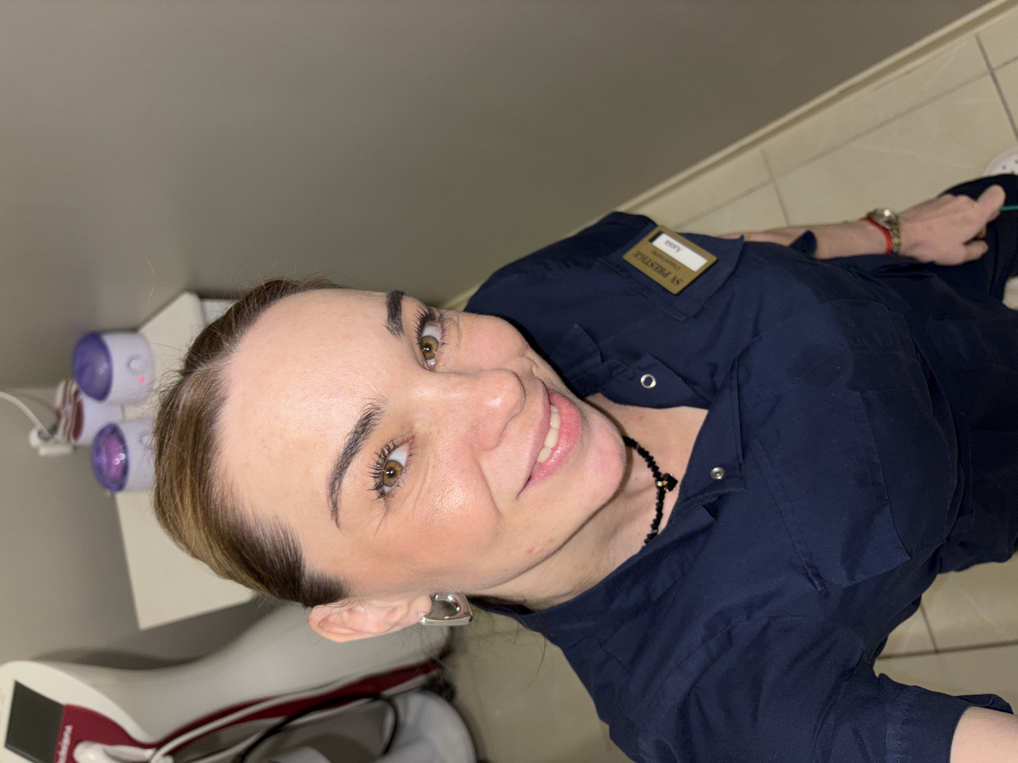 A woman is seen from a high-angle perspective, smiling and looking at the camera. She has brown hair pulled back and is wearing a navy blue shirt with a name tag that reads "SV PRESTIGE". She has a black beaded necklace, and heart-shaped earrings. A portion of her right arm is visible, showing a watch and a red string bracelet. Behind the woman and to the left are pieces of medical equipment. The background shows a neutral-colored wall and tile floor, suggesting a commercial or clinical setting. This image was taken in the city of Murino, Russia.